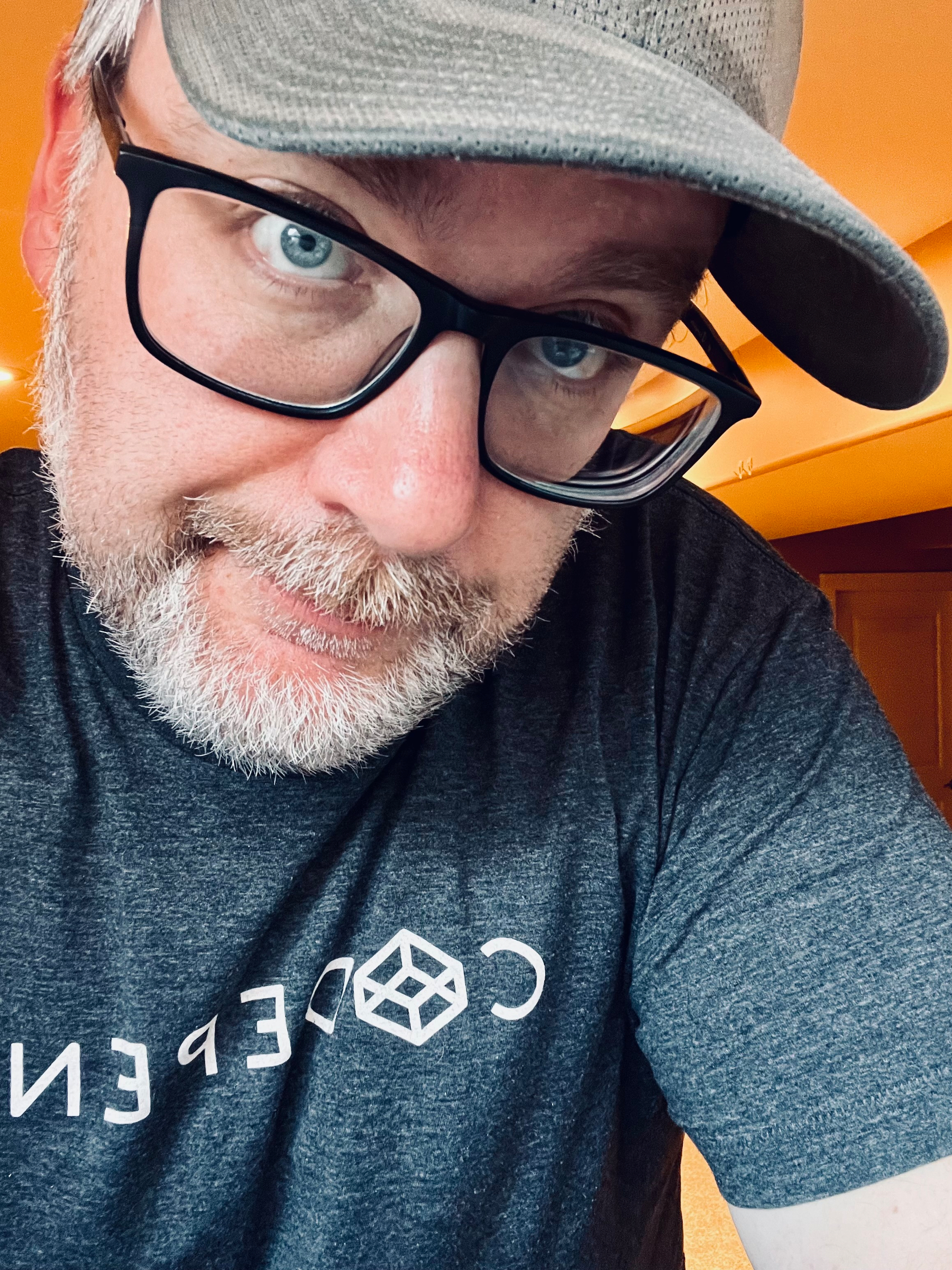 A bearded person wearing glasses and a cap is smiling slightly, dressed in a T-shirt with a geometric design.