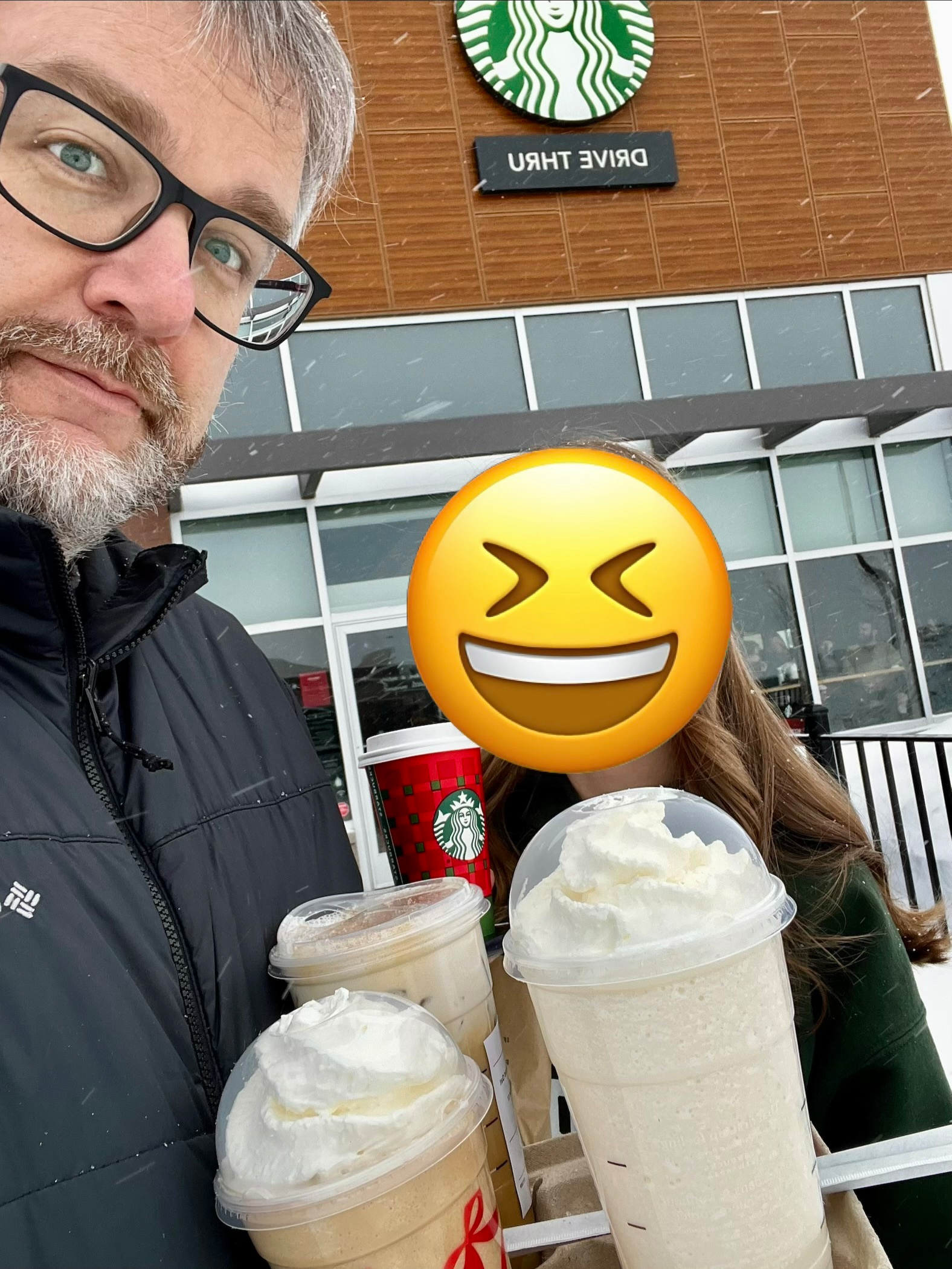 A person is holding a tray with three drinks topped with whipped cream in front of a Starbucks, accompanied by another person with a laughing emoji over their face.