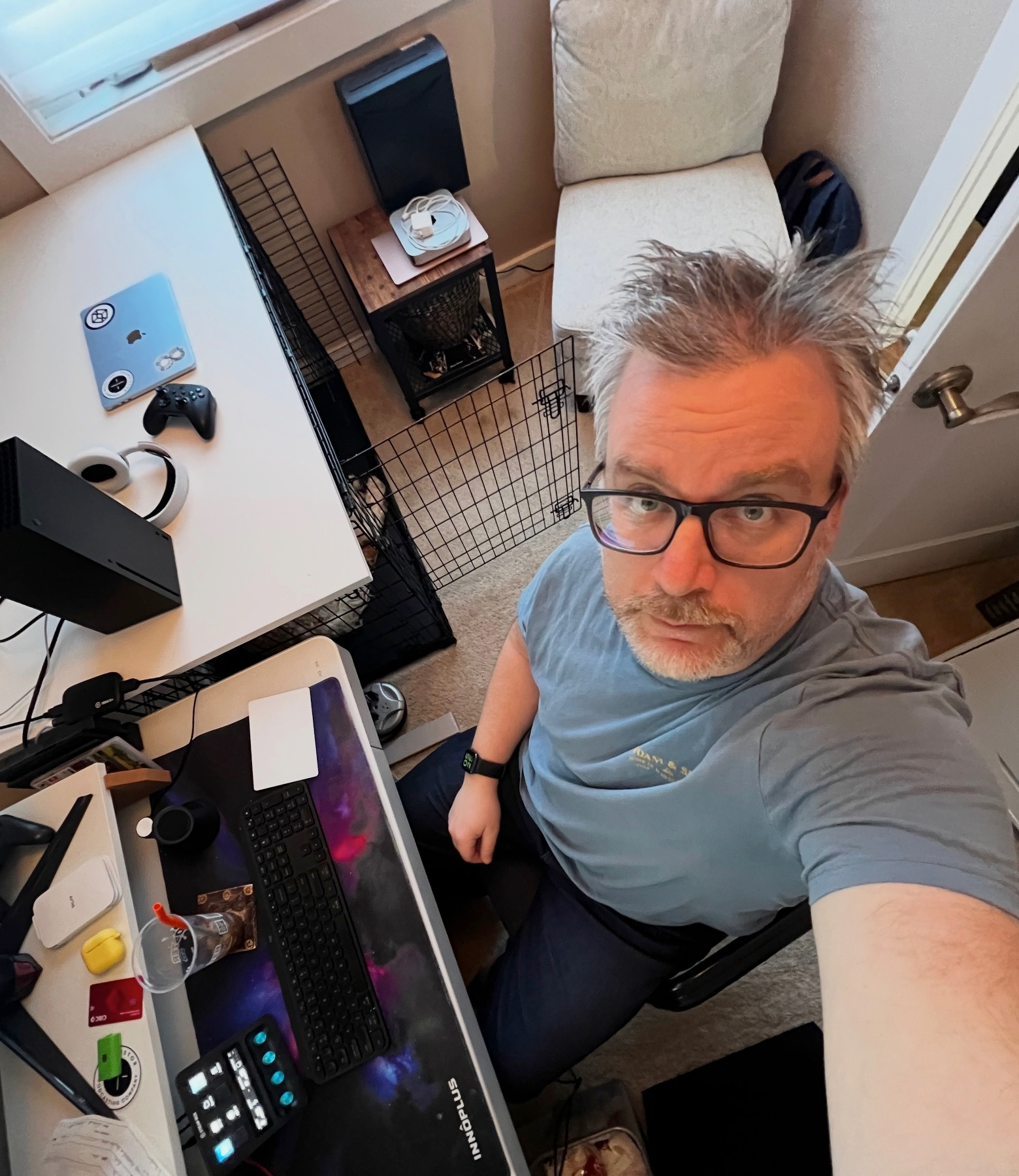 A man with glasses is taking a selfie from above in a cluttered room with a computer setup and various items around him.