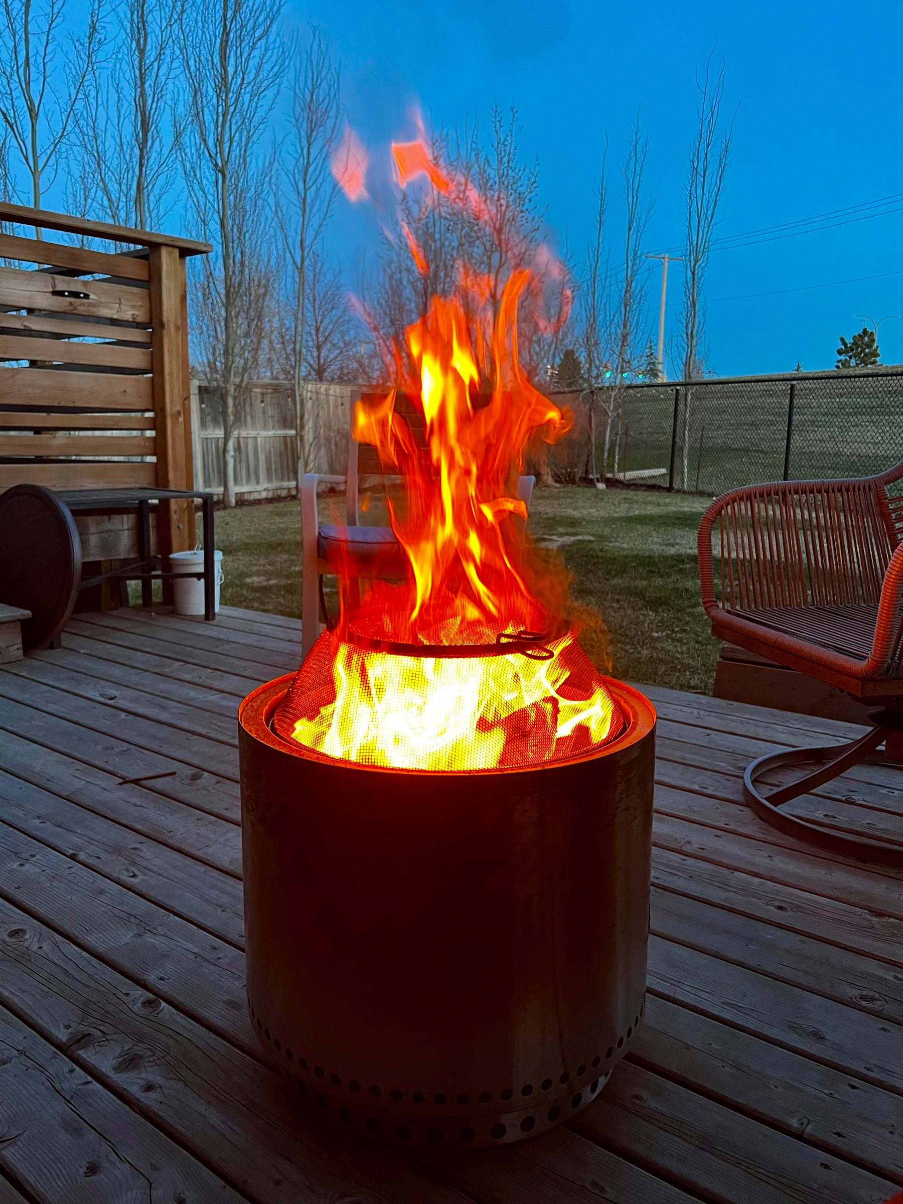 A vibrant fire burns in a metal fire pit on a wooden deck, surrounded by outdoor furniture, with a backdrop of trees and a fence.