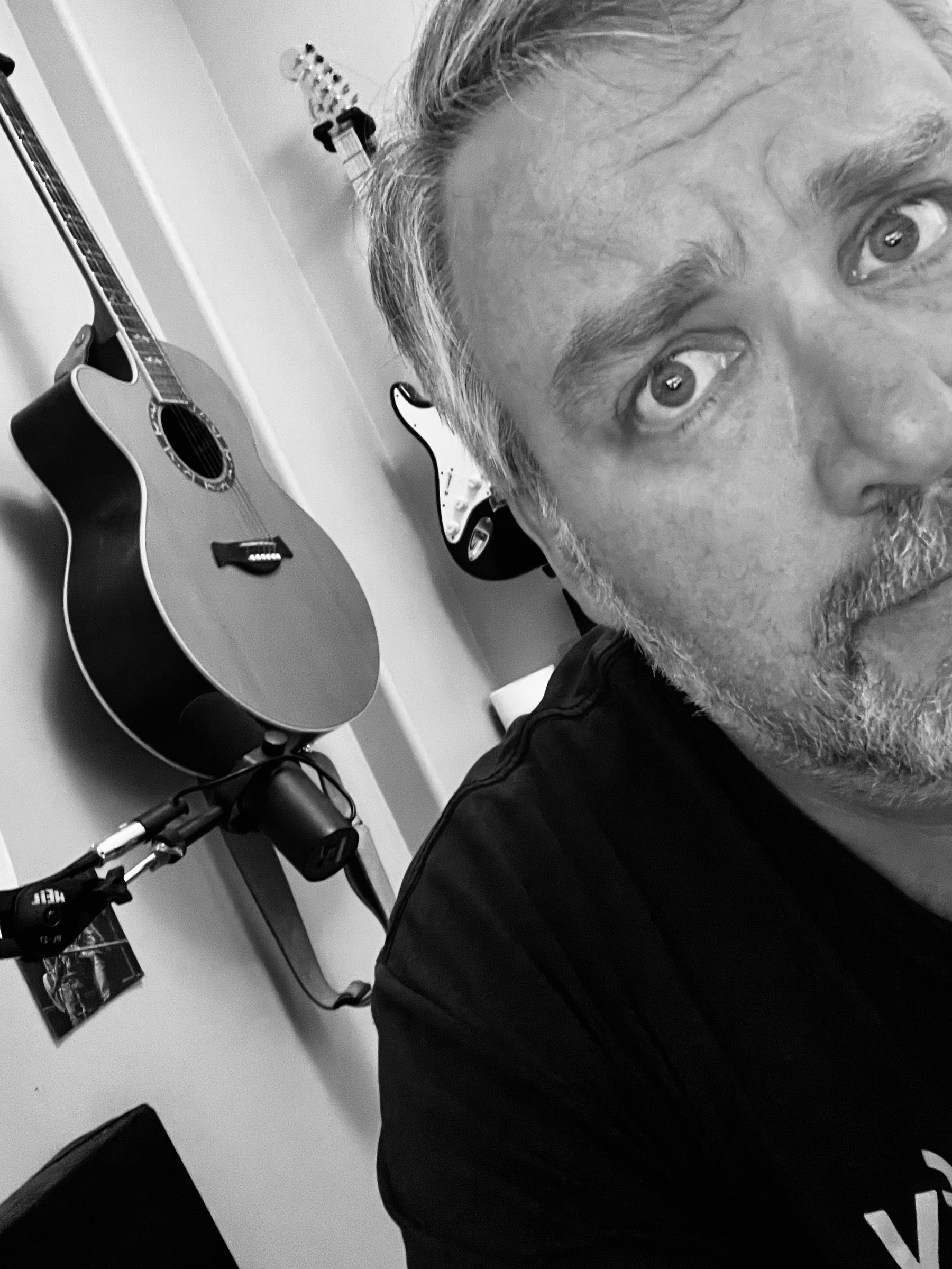 A grayscale image features a man partially in frame with a serious expression, and acoustic and electric guitars mounted on the wall behind him.