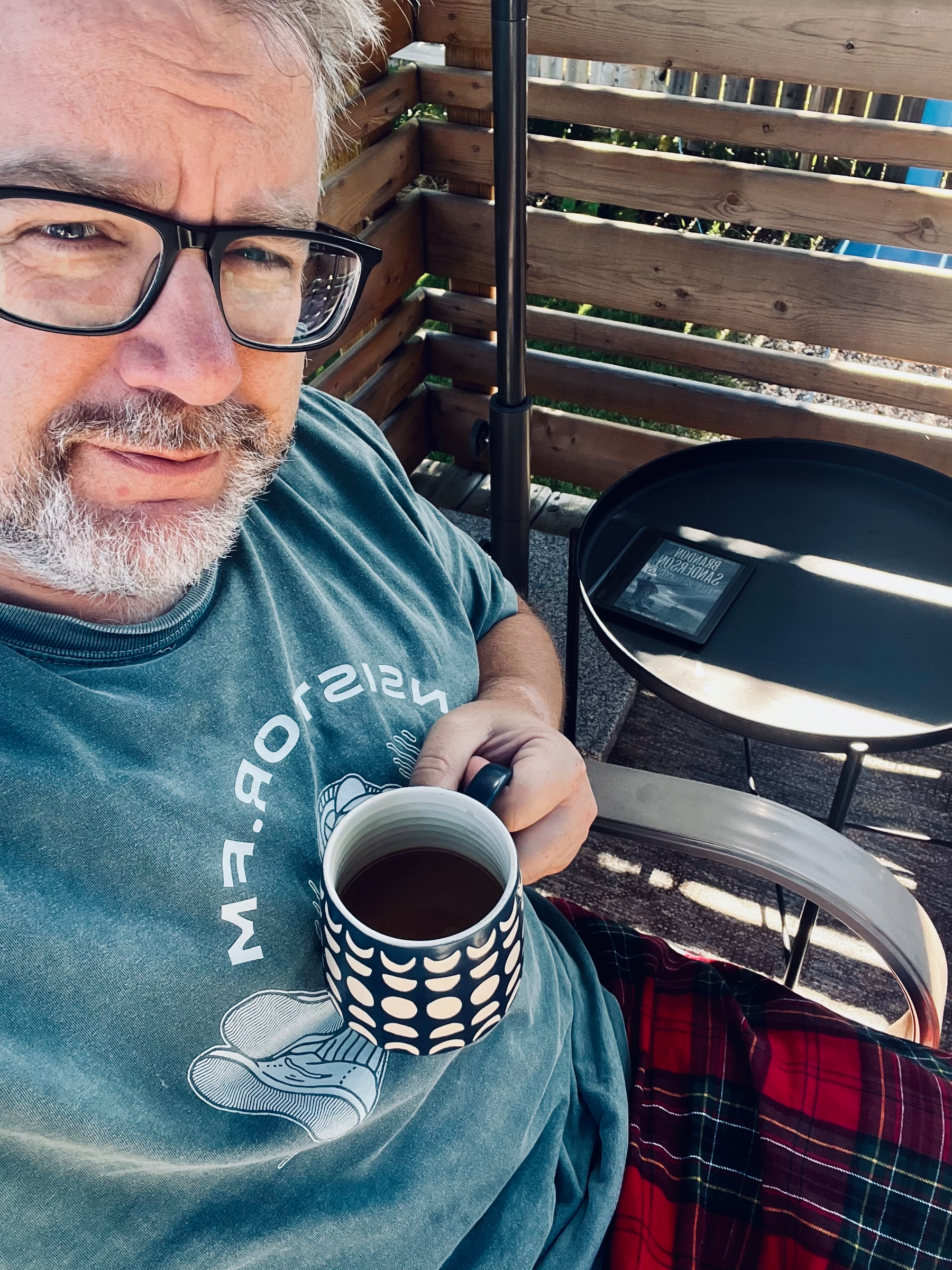 A person with glasses is sitting outdoors, holding a patterned mug, while dressed casually in a t-shirt and plaid pants.