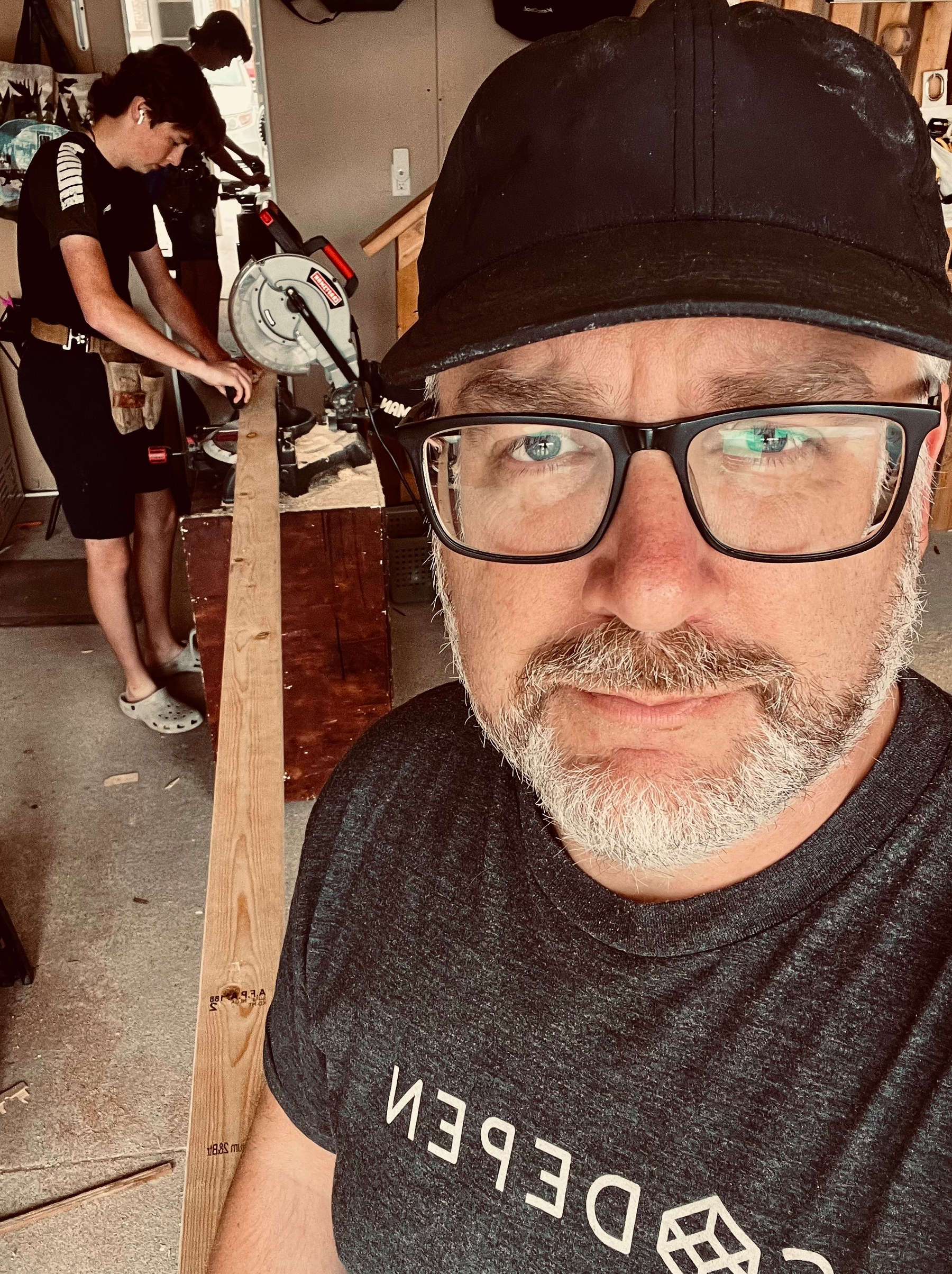 A man wearing glasses and a black cap takes a selfie while a younger person is working with a saw on a long piece of wood in a workshop.