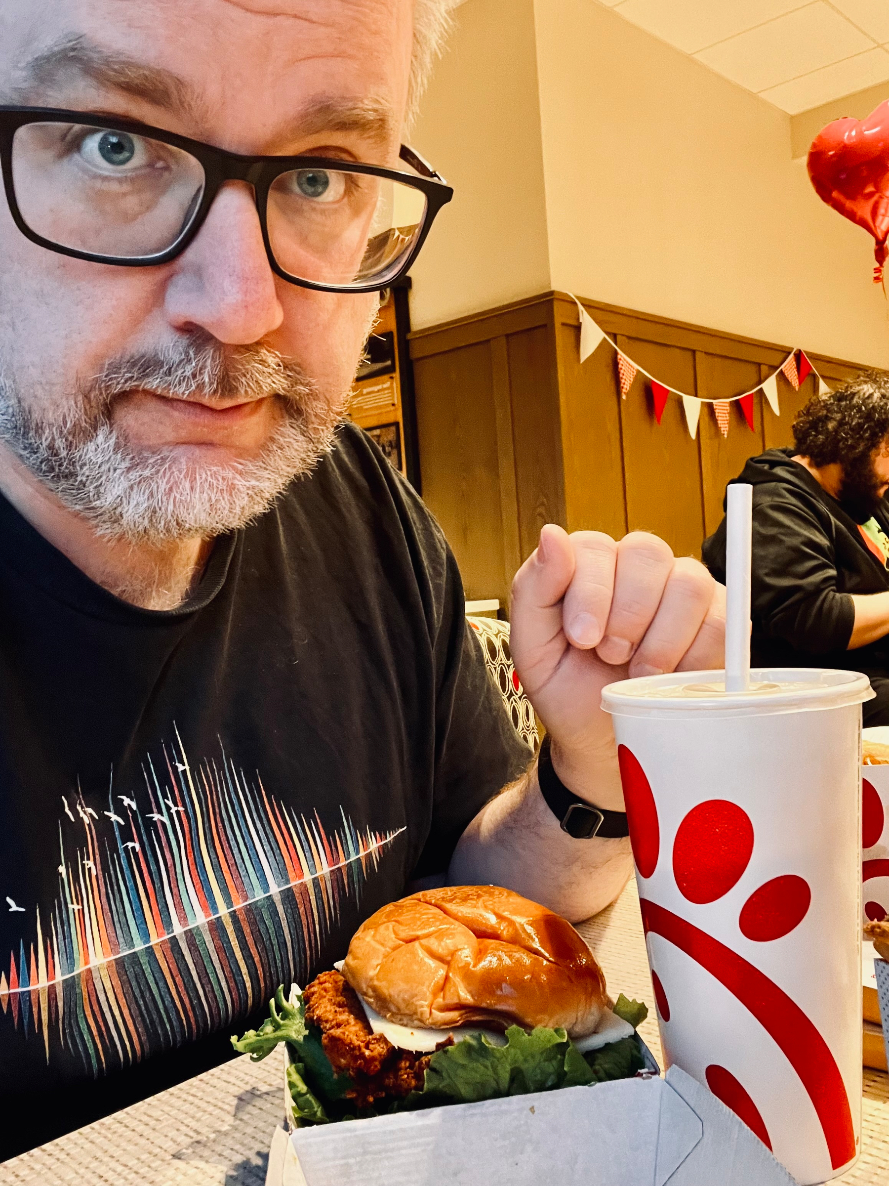 A man with glasses sits at a table with a fast-food sandwich and drink.