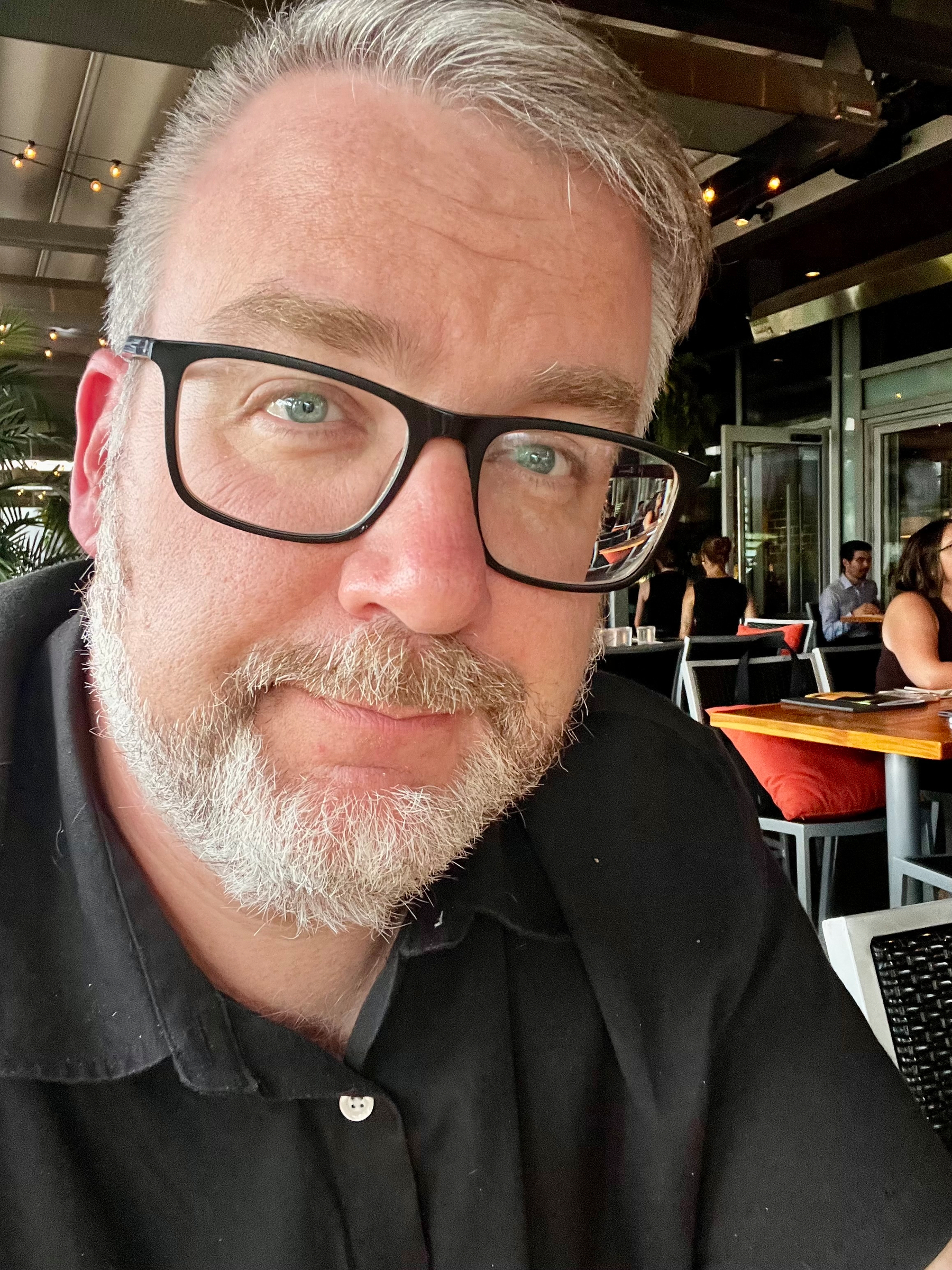 A man with gray hair, a beard, and glasses is sitting in a restaurant with other patrons visible in the background.