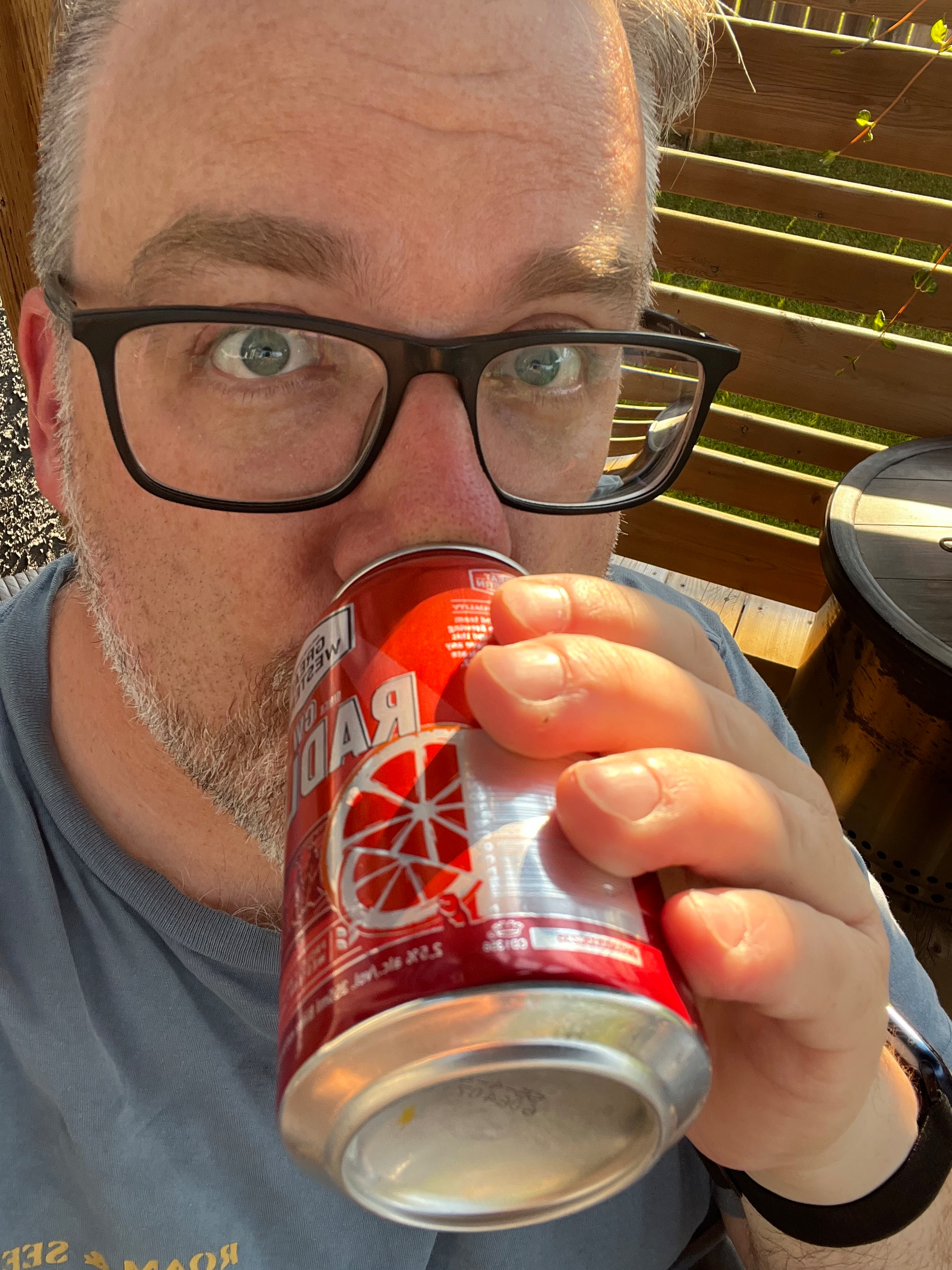 A person wearing glasses is holding and drinking from a red can with a lemon graphic on it while sitting outdoors.