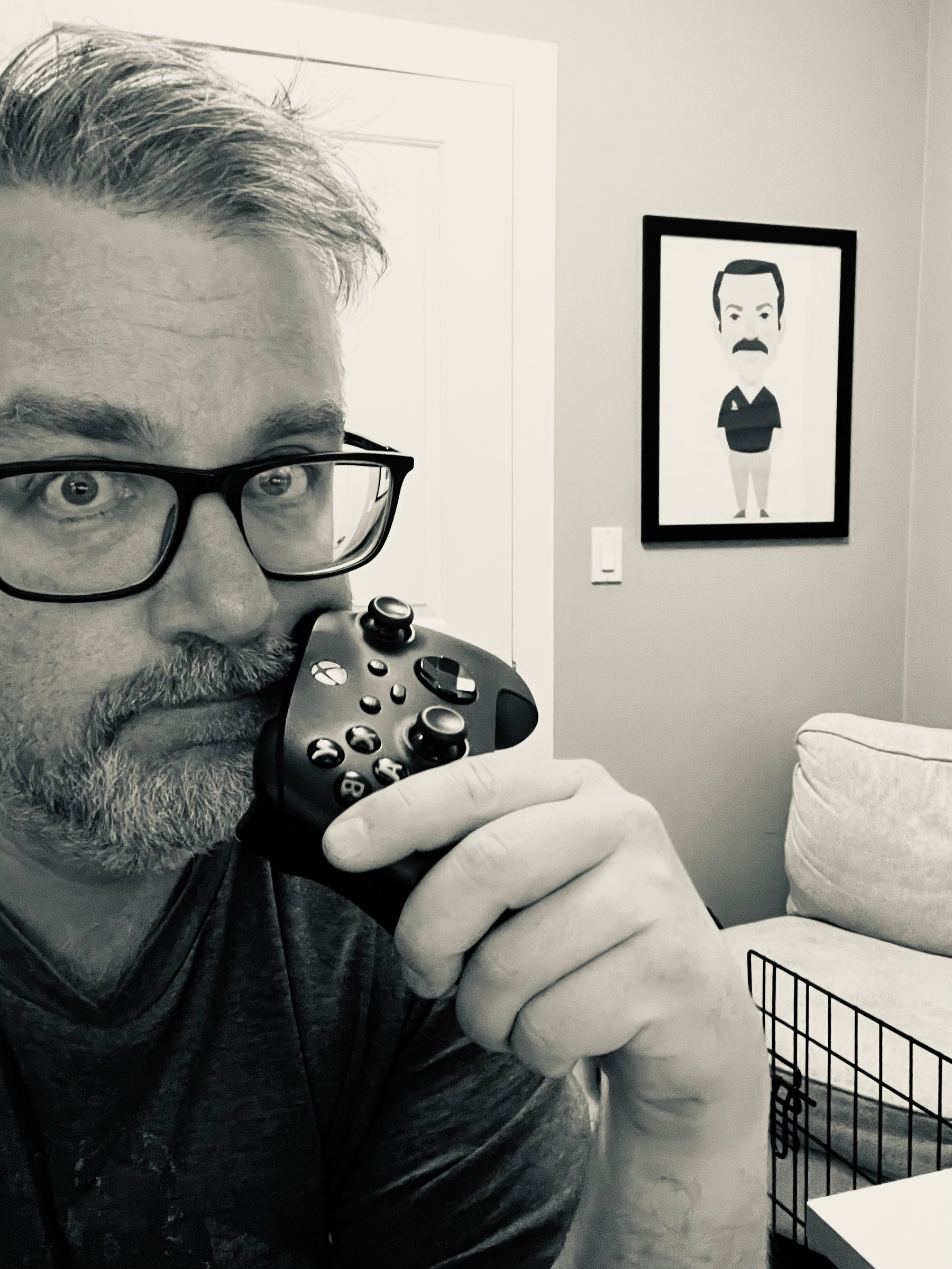 A man wearing glasses is holding a video game controller close to his face with a framed cartoon portrait of Ted Lasso on the wall behind him.