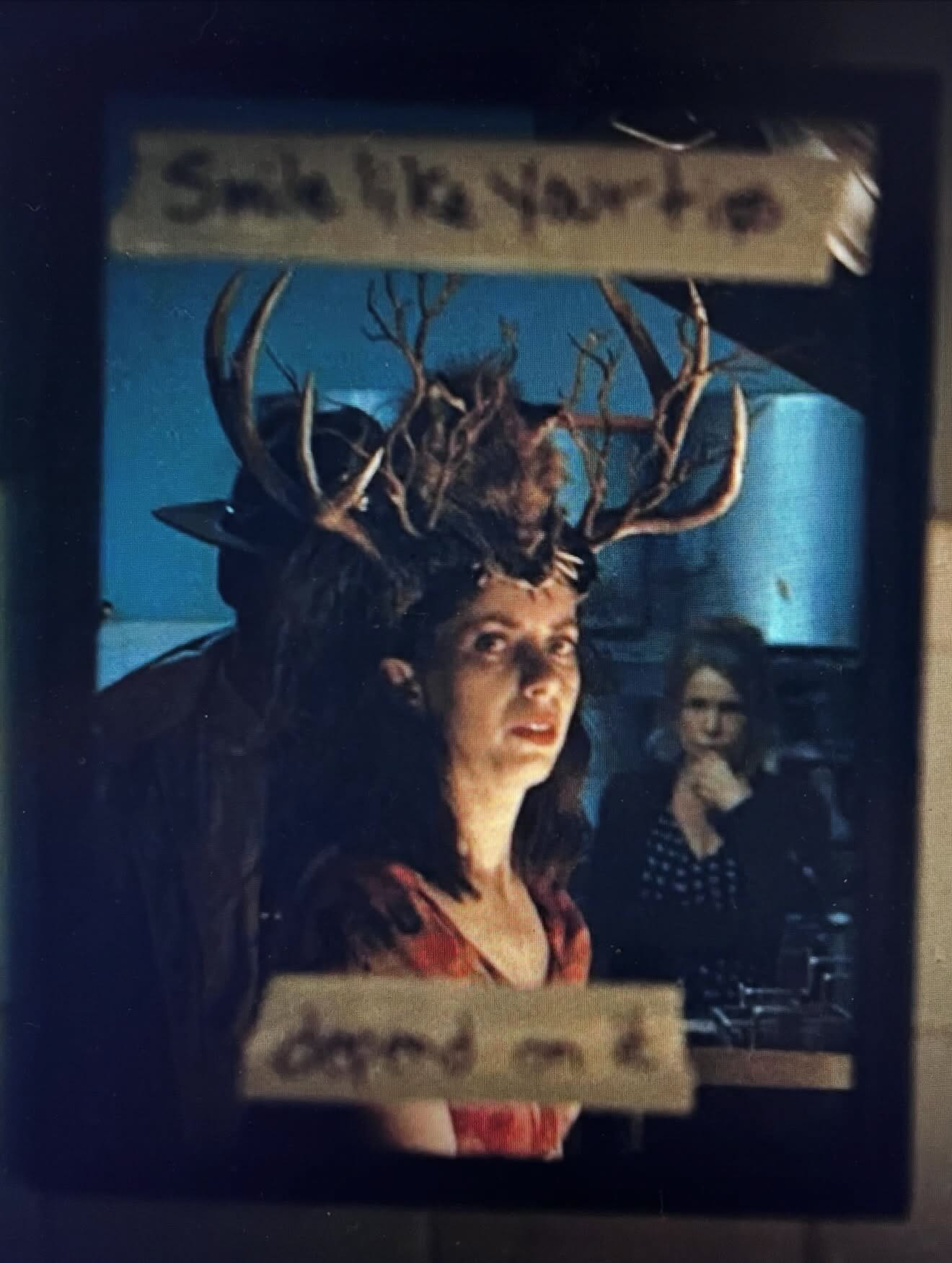 Screengrab from Widow’s Bay episode: A woman wearing an antler headpiece poses in front of a reflective surface with two people visible behind her, accompanied by taped labels saying “Smile like your tips depend on it”