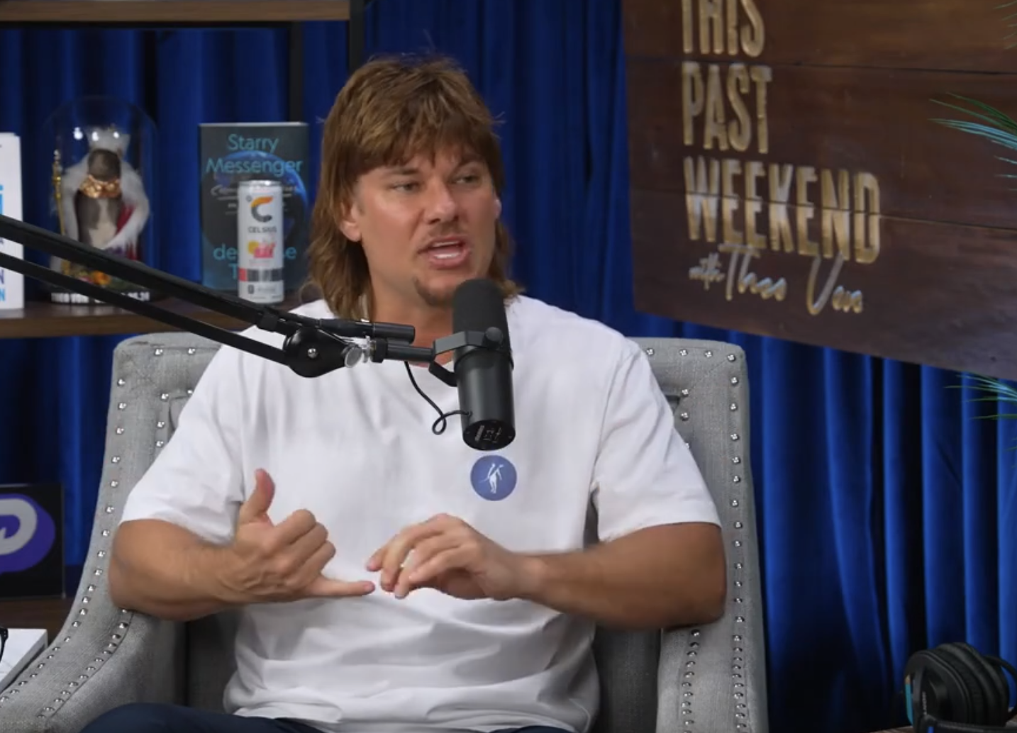 A Theo Von is speaking into a microphone on a podcast set with a sign that reads This Past Weekend in the background.