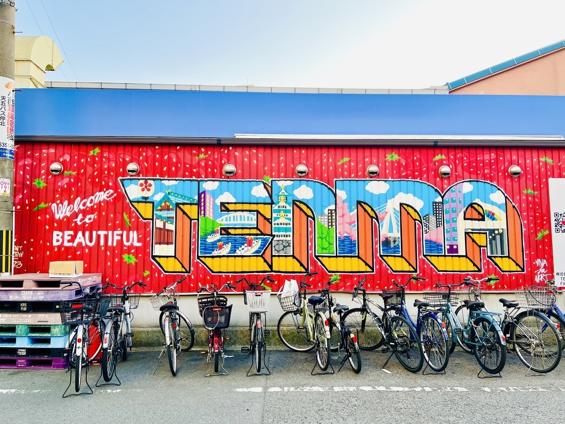 Bikes lined up along a wall with a mural that says “welcome to beautiful Tenma” in bright colours