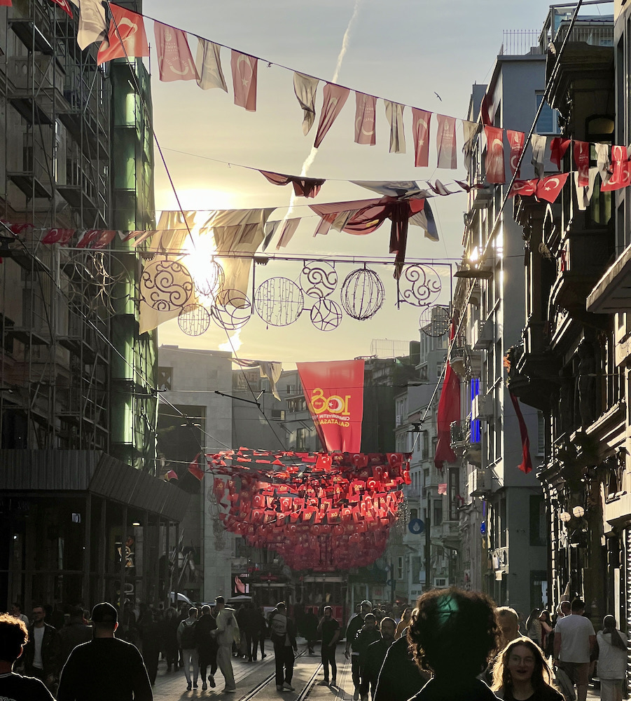 A busy shopping street with people walking. Above are strings criss-crossing the street with celebratory Turkish flags hanging down