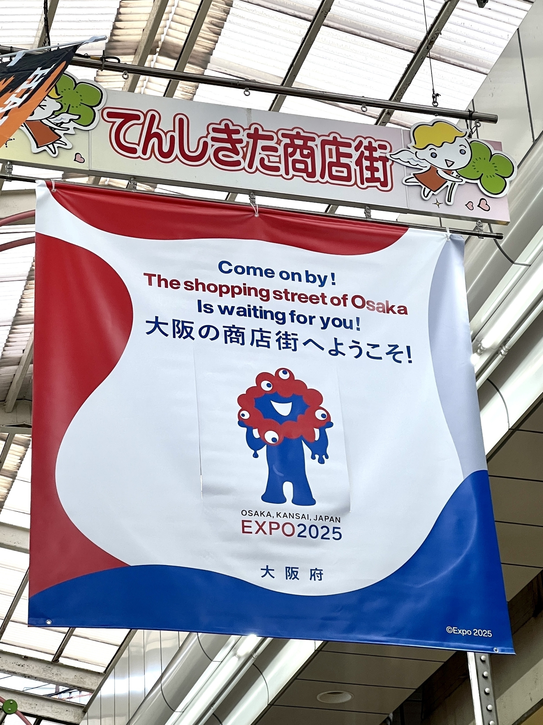 A flag hanging from the shopping arcade ceiling with Myakumyaku-kun saying: “Come on by!&10;The shopping street of Osaka&10;Is waiting for you”