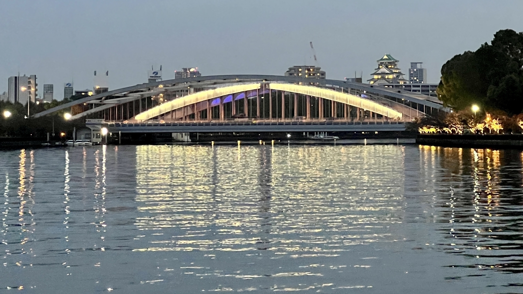 An arched bridge over the river lit up in early evening. In the background distance is Osaka Castle