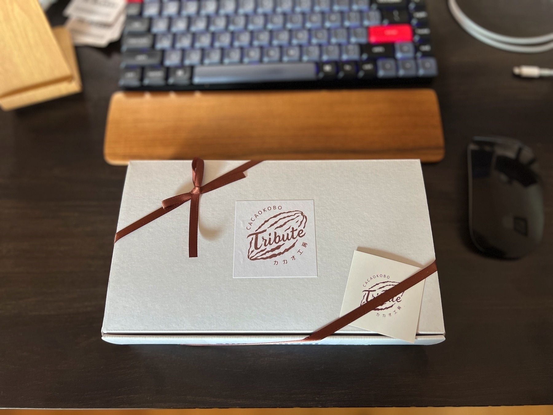 A ribboned box sits on a desk by a mechanical keyboard and mouse. The label on the box says “Cacao Kobo: Tribute”