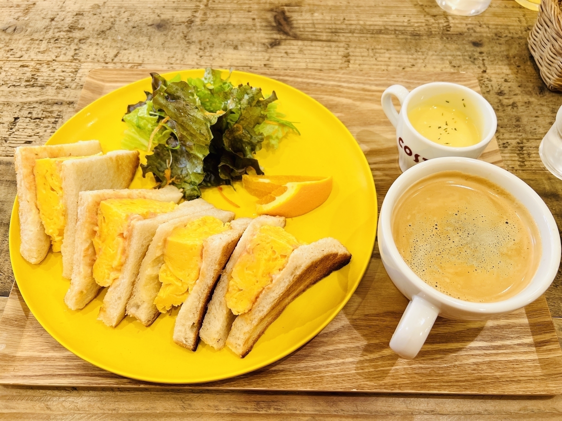 Yellow plate with egg sandwiches, green salad and an orange slice, beside is a cup of yellow corn soup, and a cup of coffee
