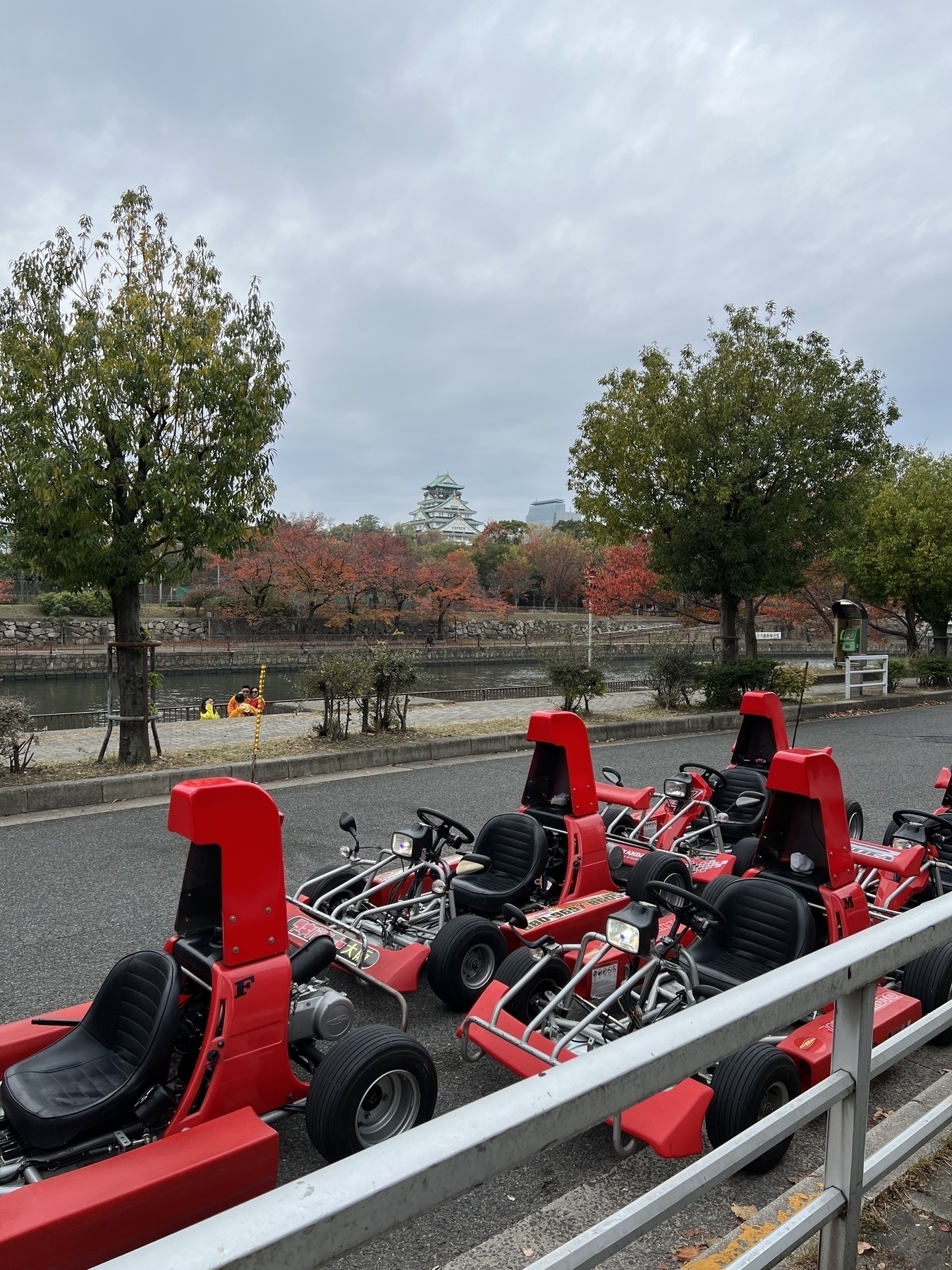 Five gokarts are parked on the side of the road. Drivers dressed in costumes (like Winnie the Pooh) are down by the moat taking photos with Osaka Castle in the background