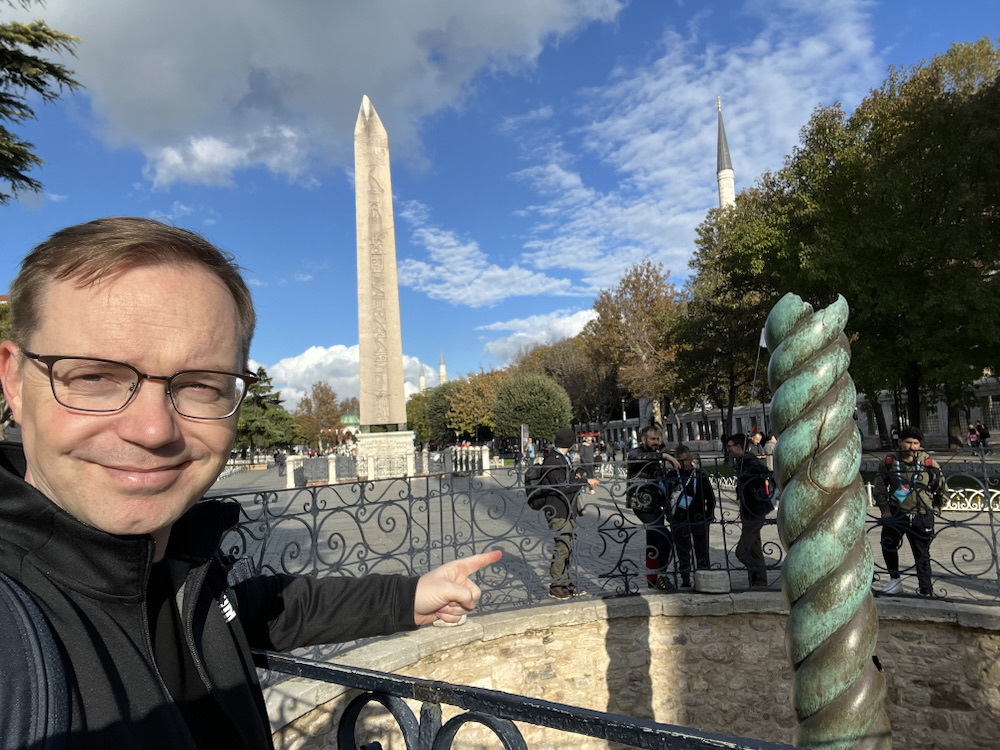 Chad taking a selfie of him pointing at the Serpent Column. In the background is an Egyptian obelisk