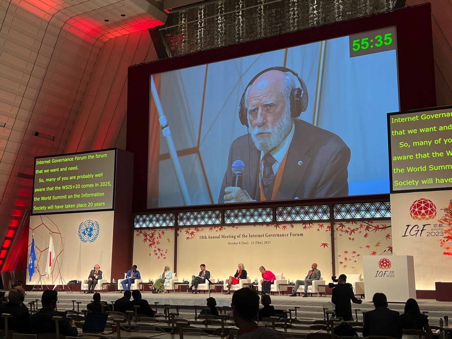 A stage with 8 speakers. A large screen above displays the face of Vint Cerf who is speaking