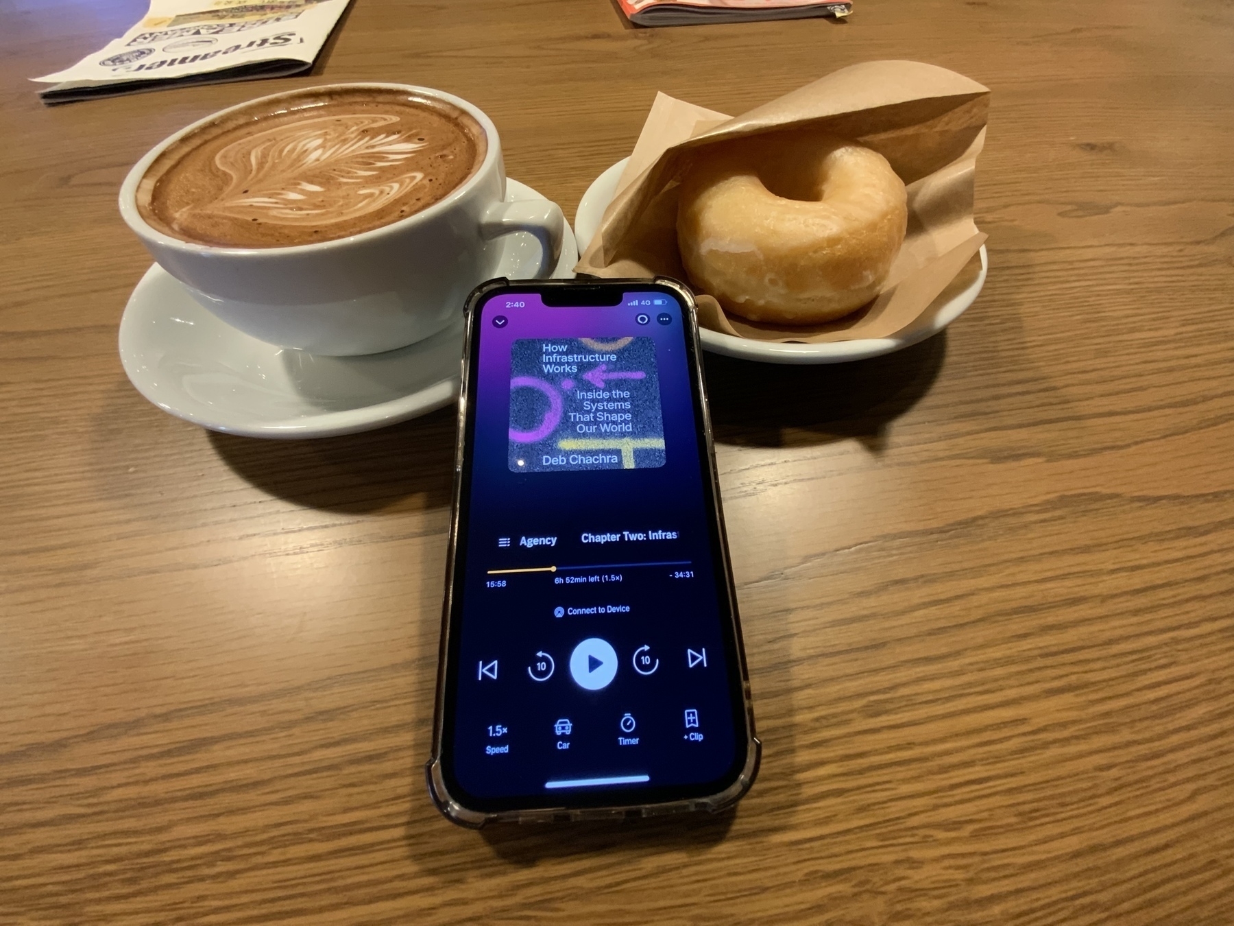 iPhone with the book playing on it resting against a mug of mocha and a plate with a donut