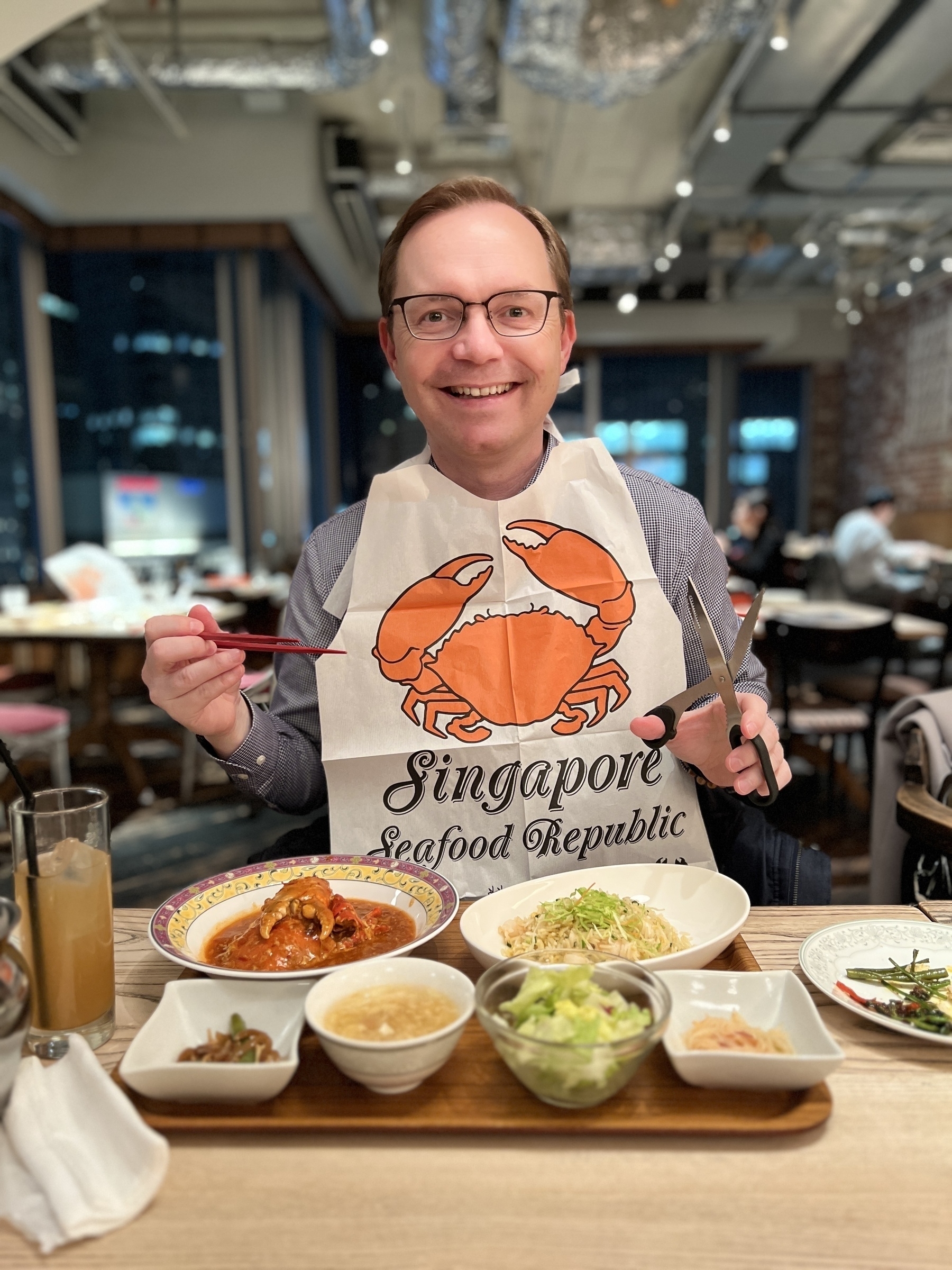 Chad in a restaurant with a plate full of food in front of him wearing a paper bib with the logo for “Singapore Seafood Republic” brandishing chopsticks and a pair of scissors and looking like a happy psycho
