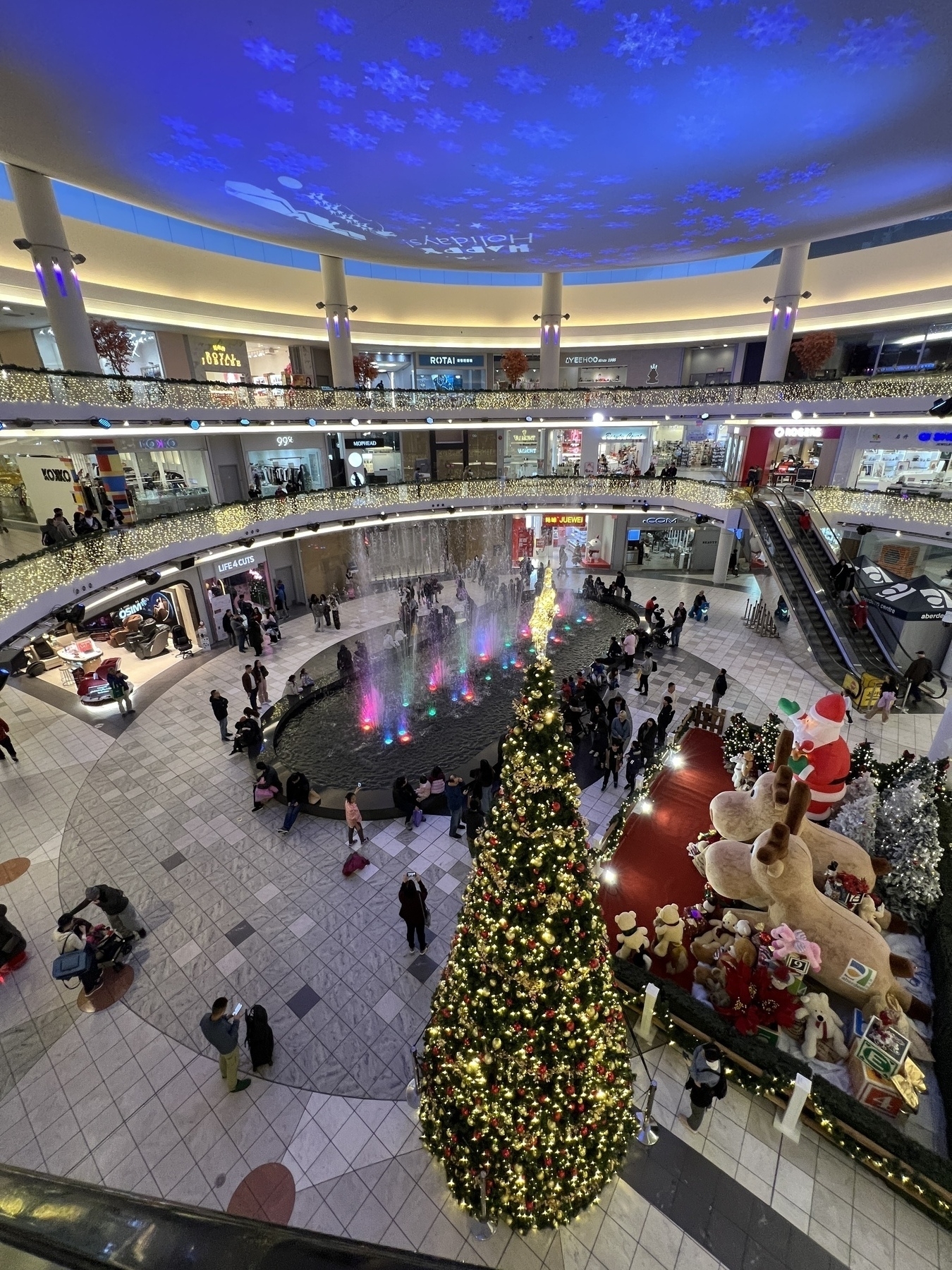 From the top floor of a shopping mall looking into a plaza where a fountain show is happening. There is a giant Xmas tree and a huge plush reindeer and Santa display