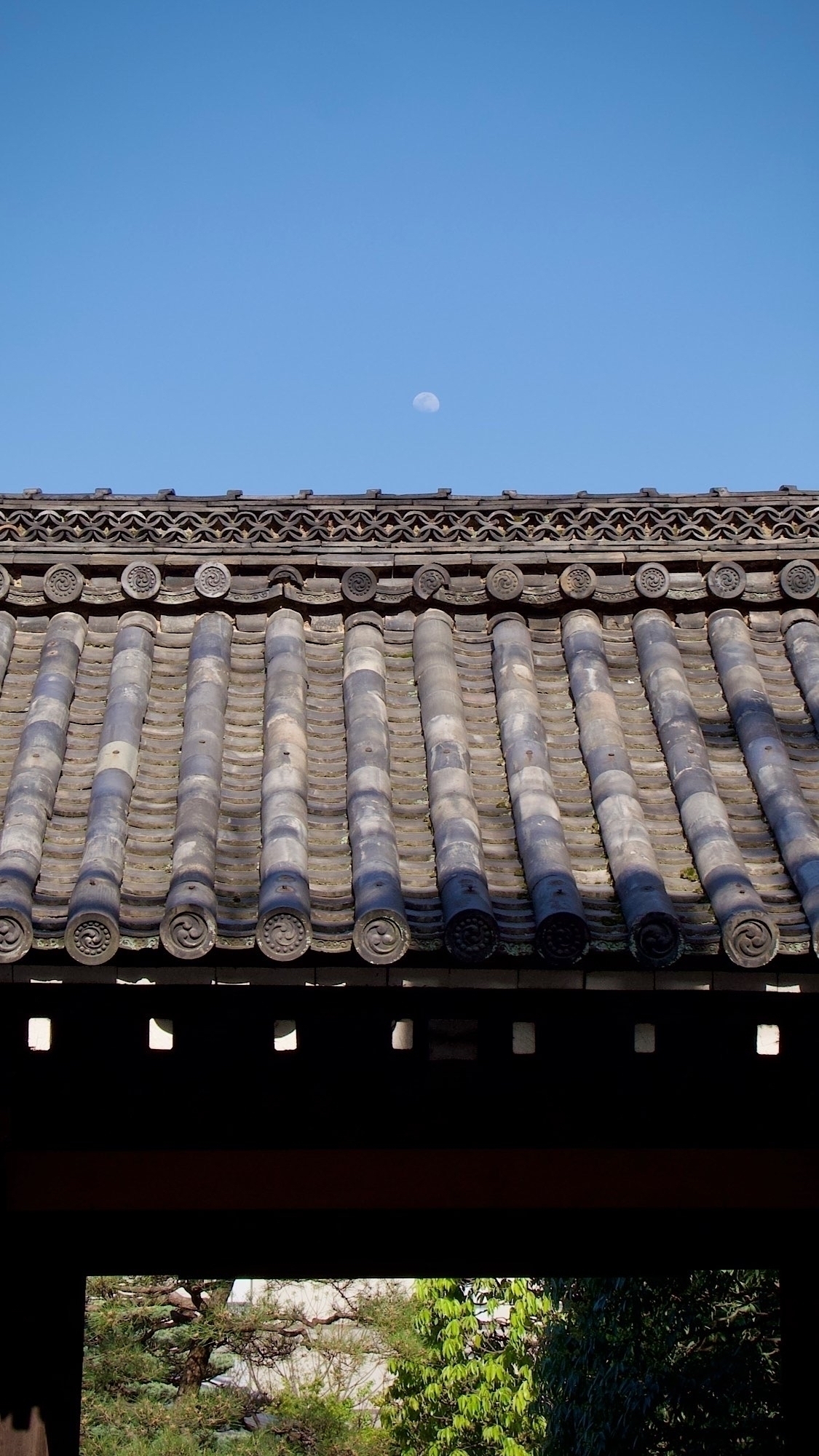 Above the roof of a temple gate the moon is visible in a perfectly clear blue sky