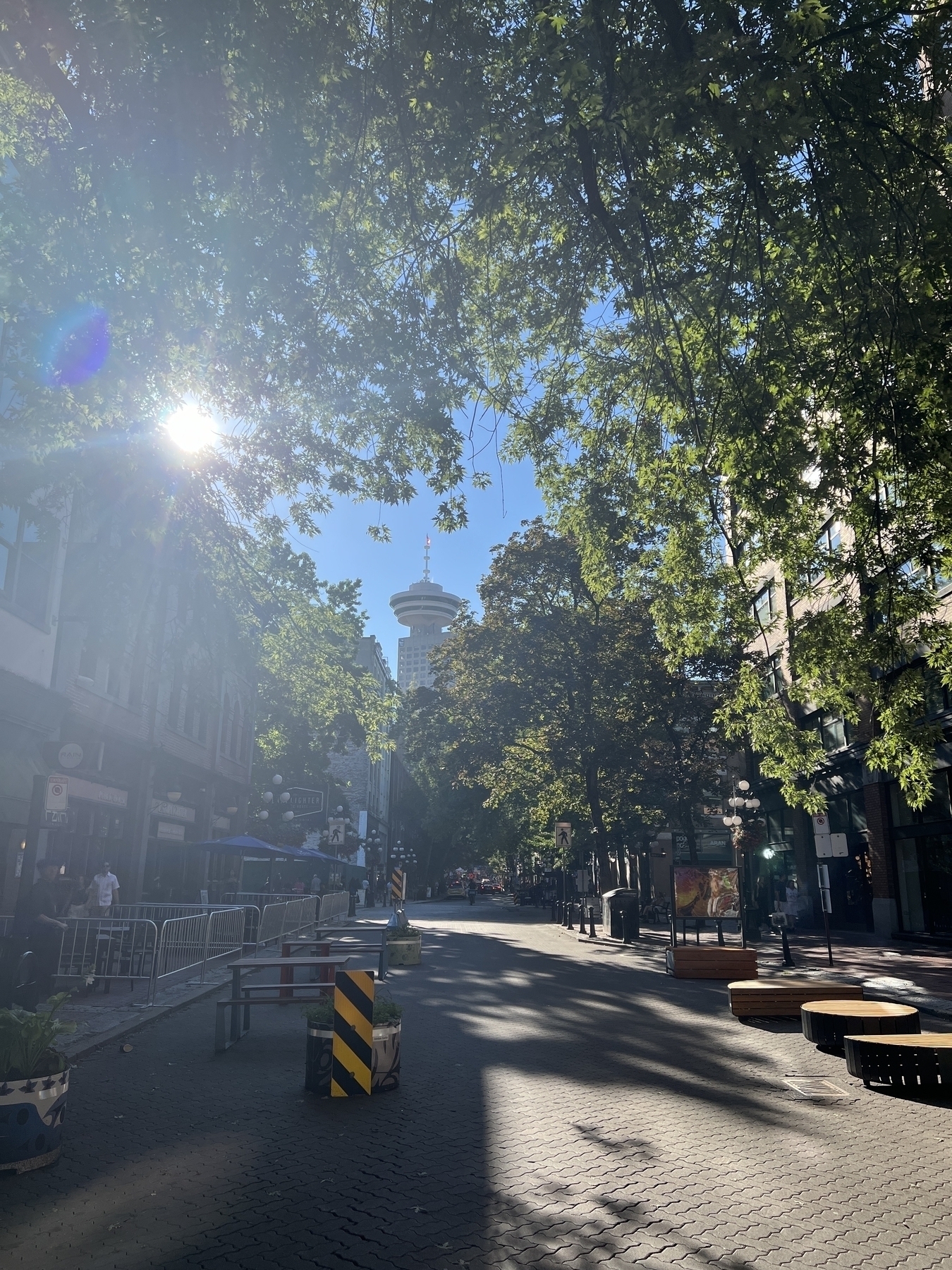 Water street in Gastown. The sun shines through the leaves of overhanging trees, Harbour Center visible in the distance