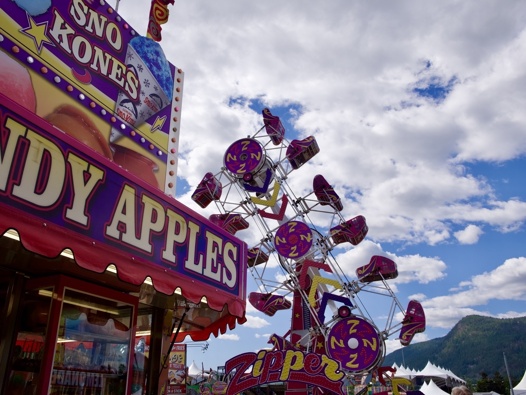 A carnival stall selling Sno Kknes and Candy Apples, in the distance is the Zipper Carnival Ride