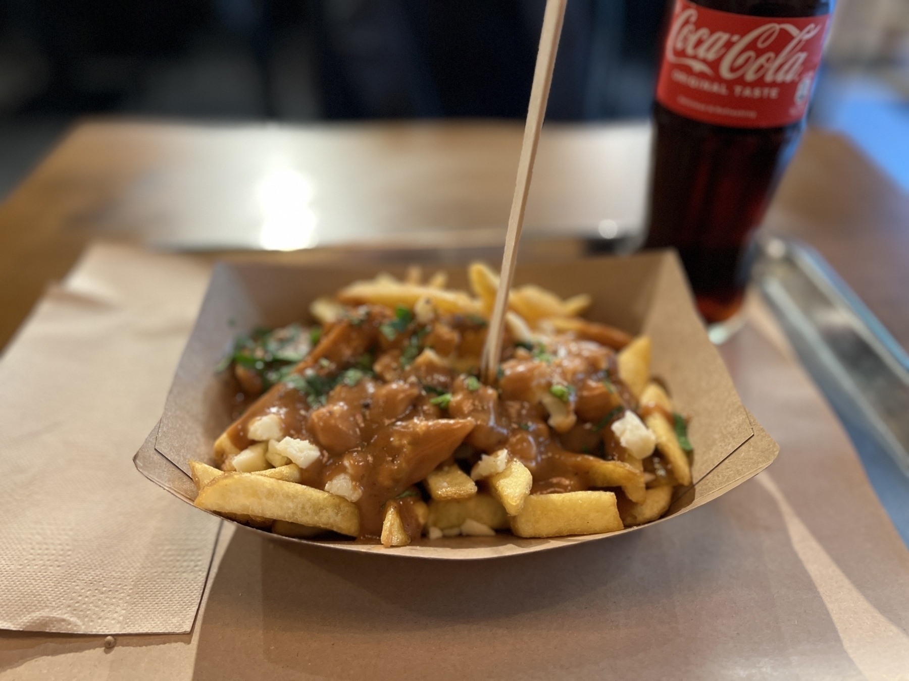 Paper plate of poutine with a wooden disposable fork stuck in it. A glass bottle of coke in the background 