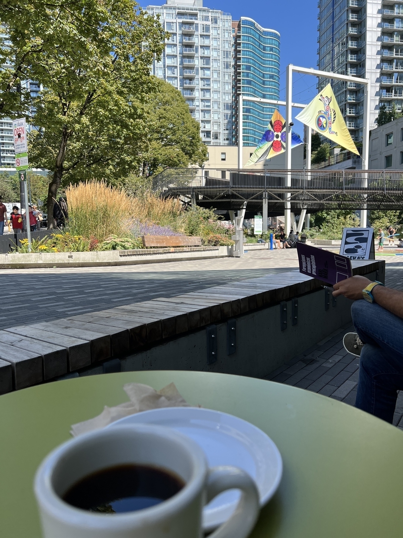 Tight shot of the top of a mug of coffee, the arm of the man next to me holding “Formaciones económicas precapitalistas” with a public park in the background featuring large shade sails overhead with indigenous art