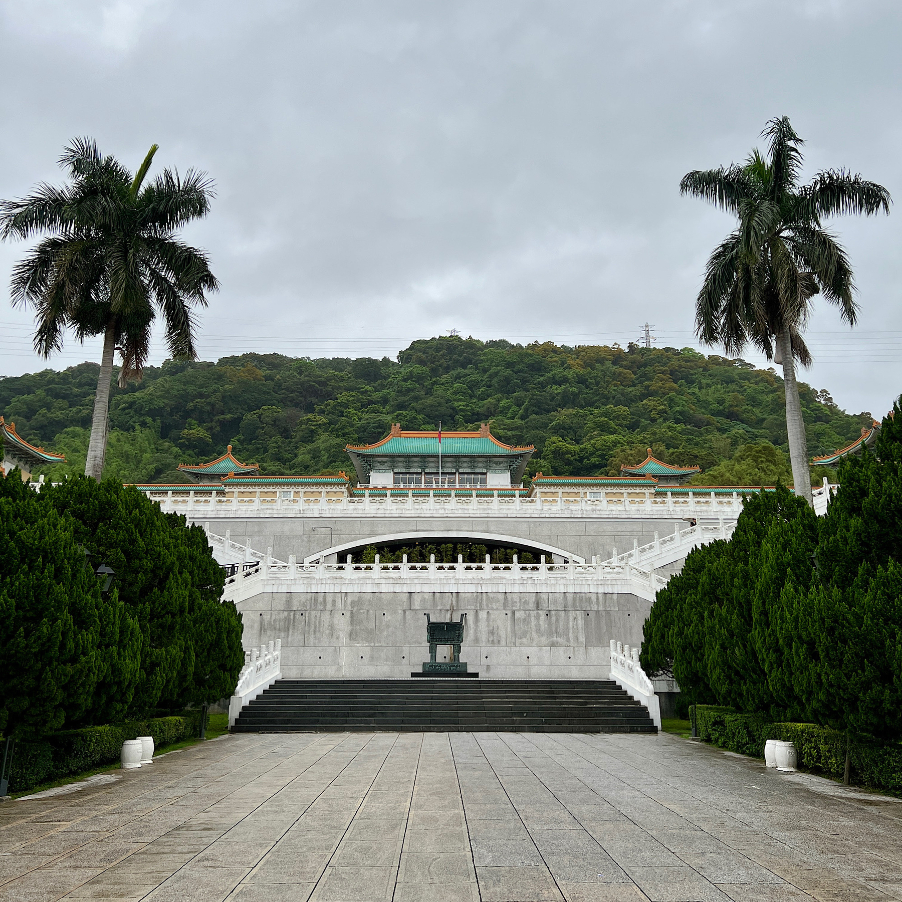Path leading up to the National Palace Museum in Taipei, a white building backed by a green mountain and flanked by palm trees