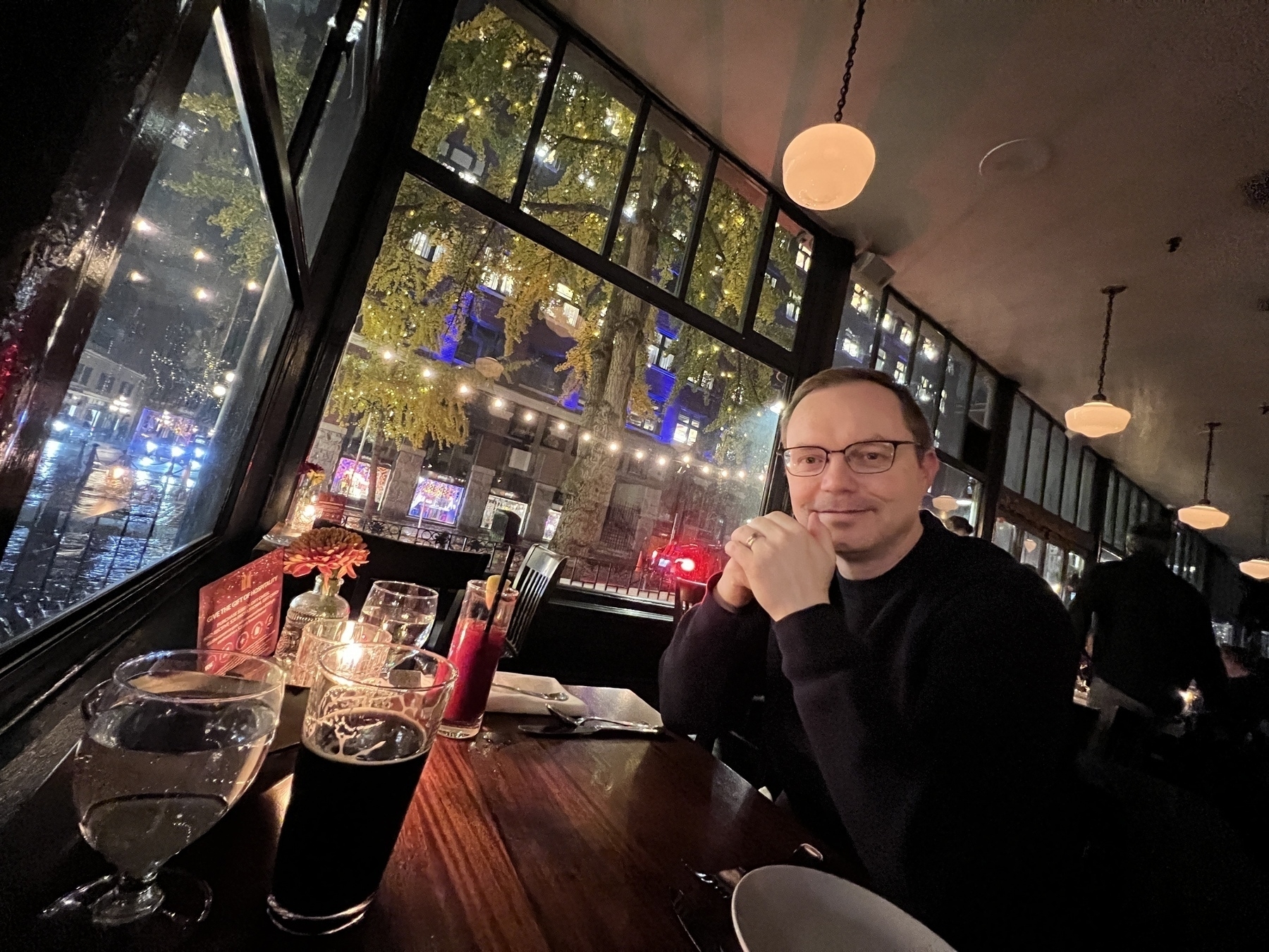 Wide angle shot of Chad sitting at a low lit dining table. In front of him are large windows opening onto a well lit rainy street