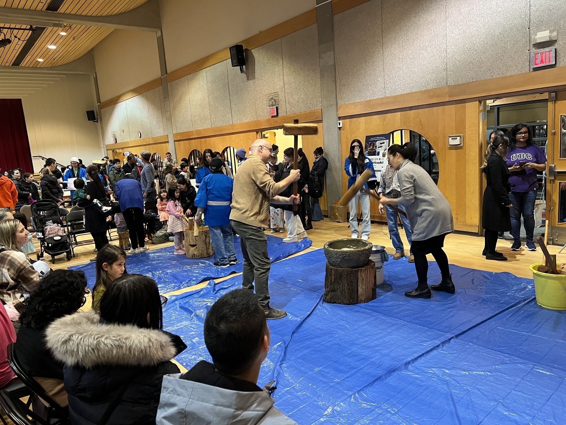 People with large wooden mallets taking turns hammering mocha in large stone bowl. The floor is covered in blue tarp, and people sit around the edges watching and waiting their turn