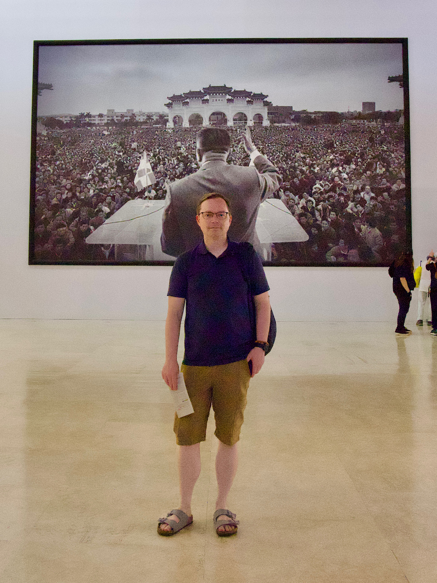 Chad standing in front of an epic and very large photo of a 1990 pro-democracy protest image of a speaker from behind exhorting a crowd of students 