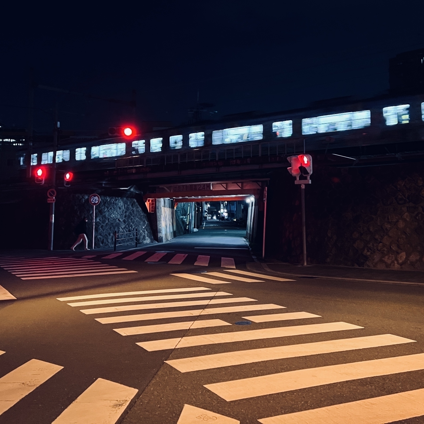 Late night photo. Train passes by. Below is a short tunnel under the tracks, with someone about to walk through. The foreground shows an empty crosswalk scramble. 