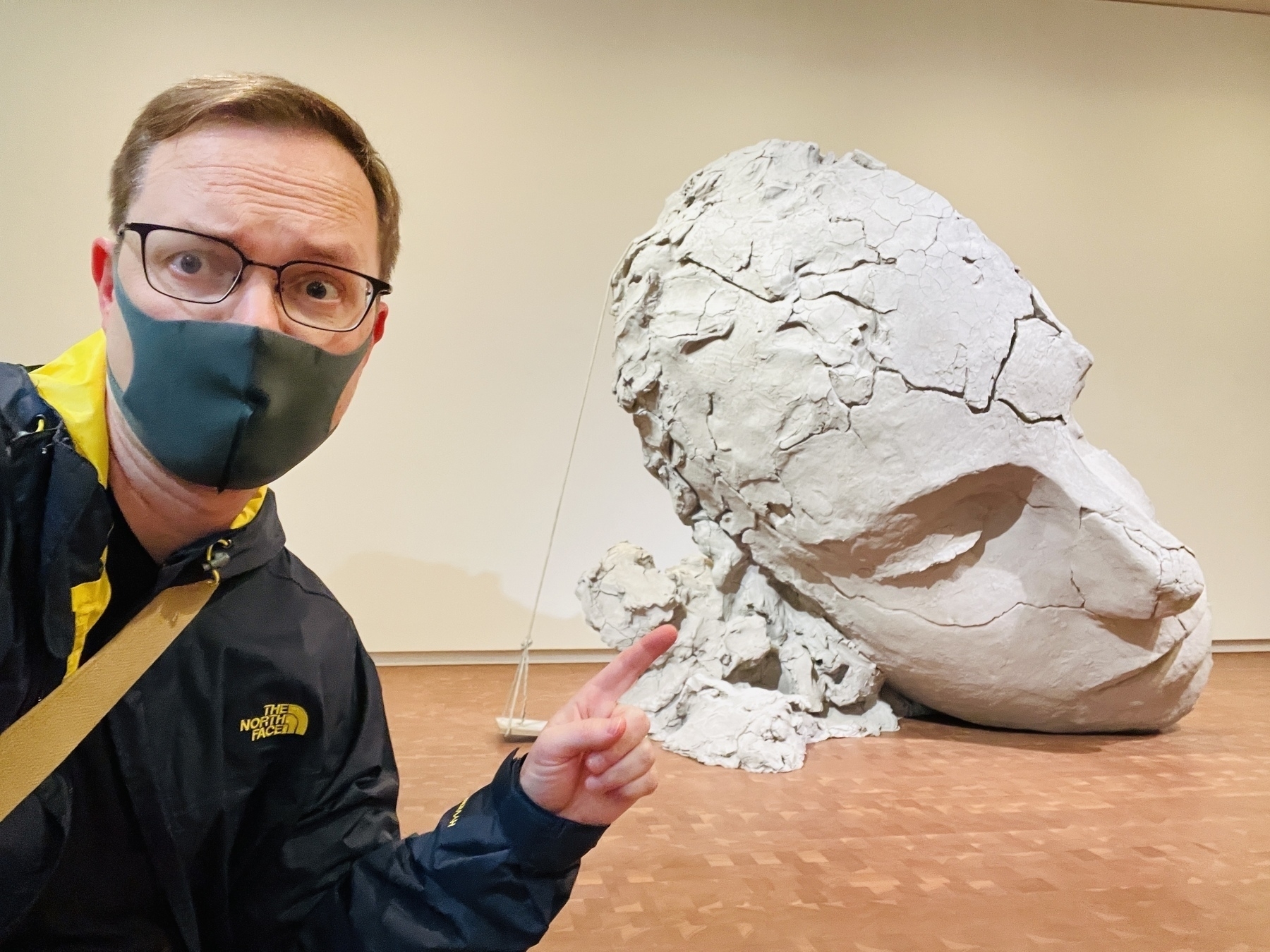 Chad selfie in front of a 5 foot tall sculpture of half a head in what looks like dry cracked clay, and positioned like it has fallen on the floor