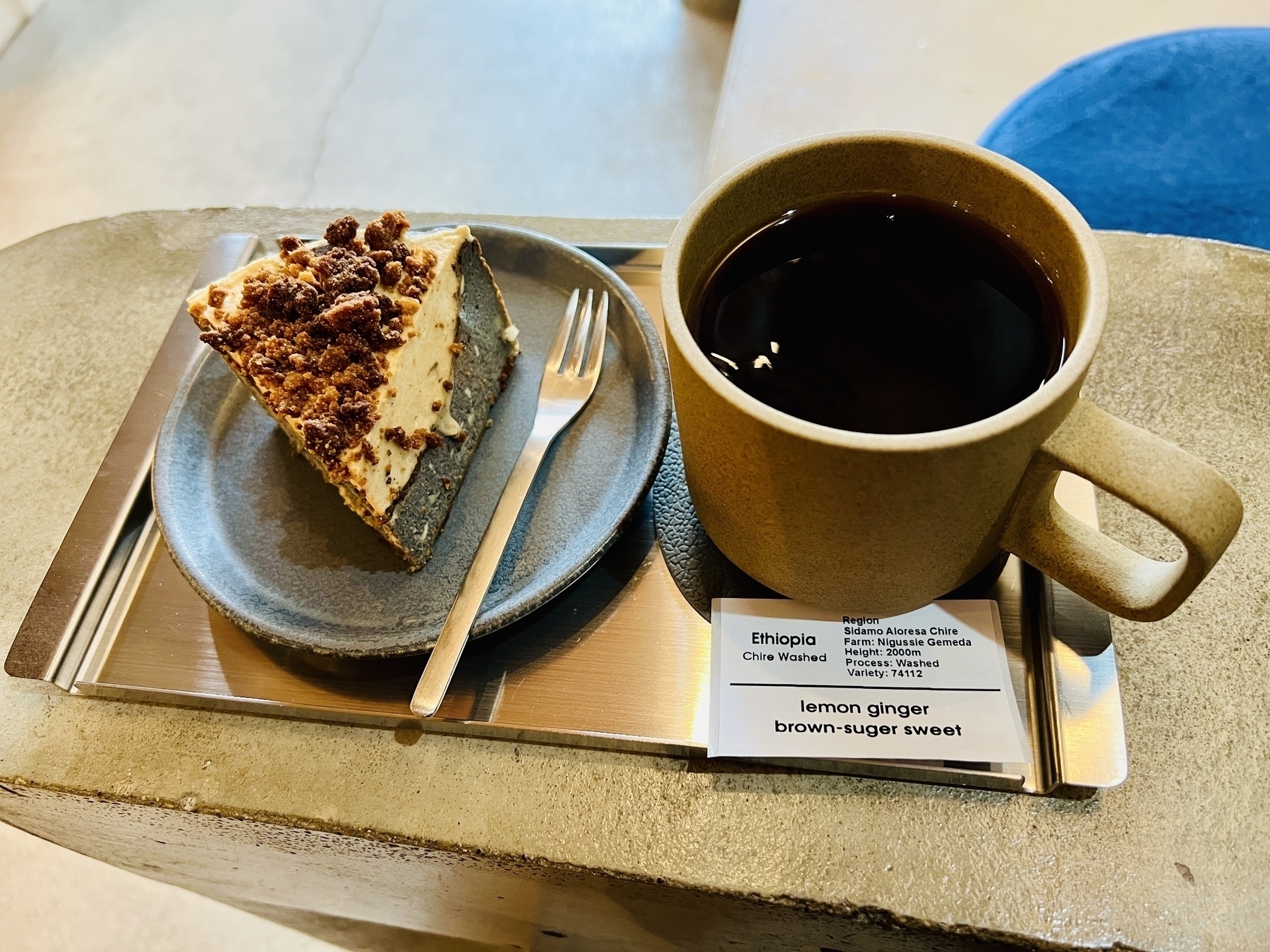 Close shot of a slice of cheesecake and a cup of black coffee. A paper label describes the coffee: &10;&10;Ethiopia&10;Chire Washed&10;Region&10;Sidamo Aloresa Chire&10;Farm: Nigussie Gemeda&10;Height: 2000m&10;Process: Washed&10;Variety: 74112&10;lemon ginger brown-suger sweet