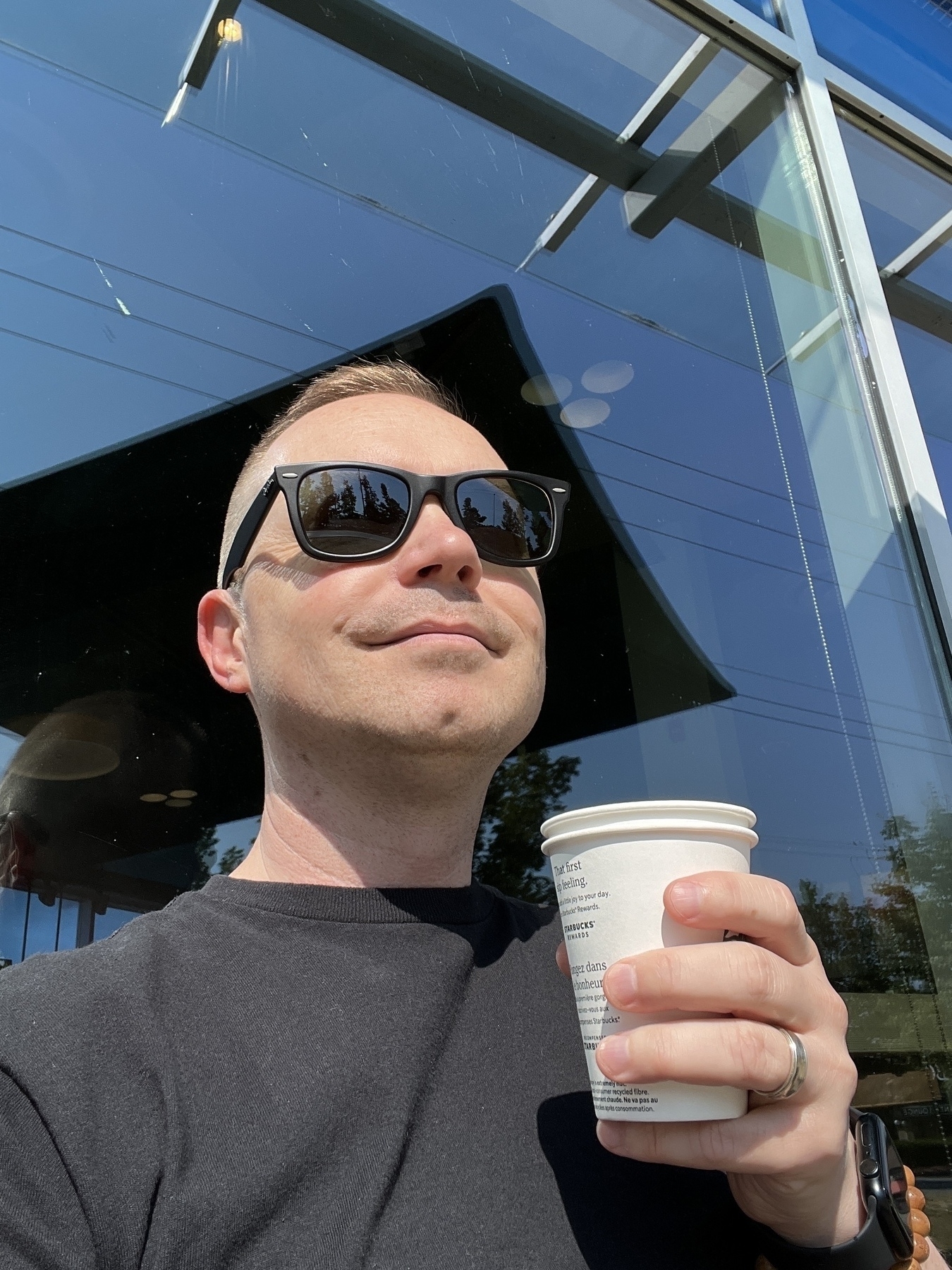 Chad selfie sitting on a cafe patio looking up at the clear sky with sunglasses on, holding a paper cup of coffee