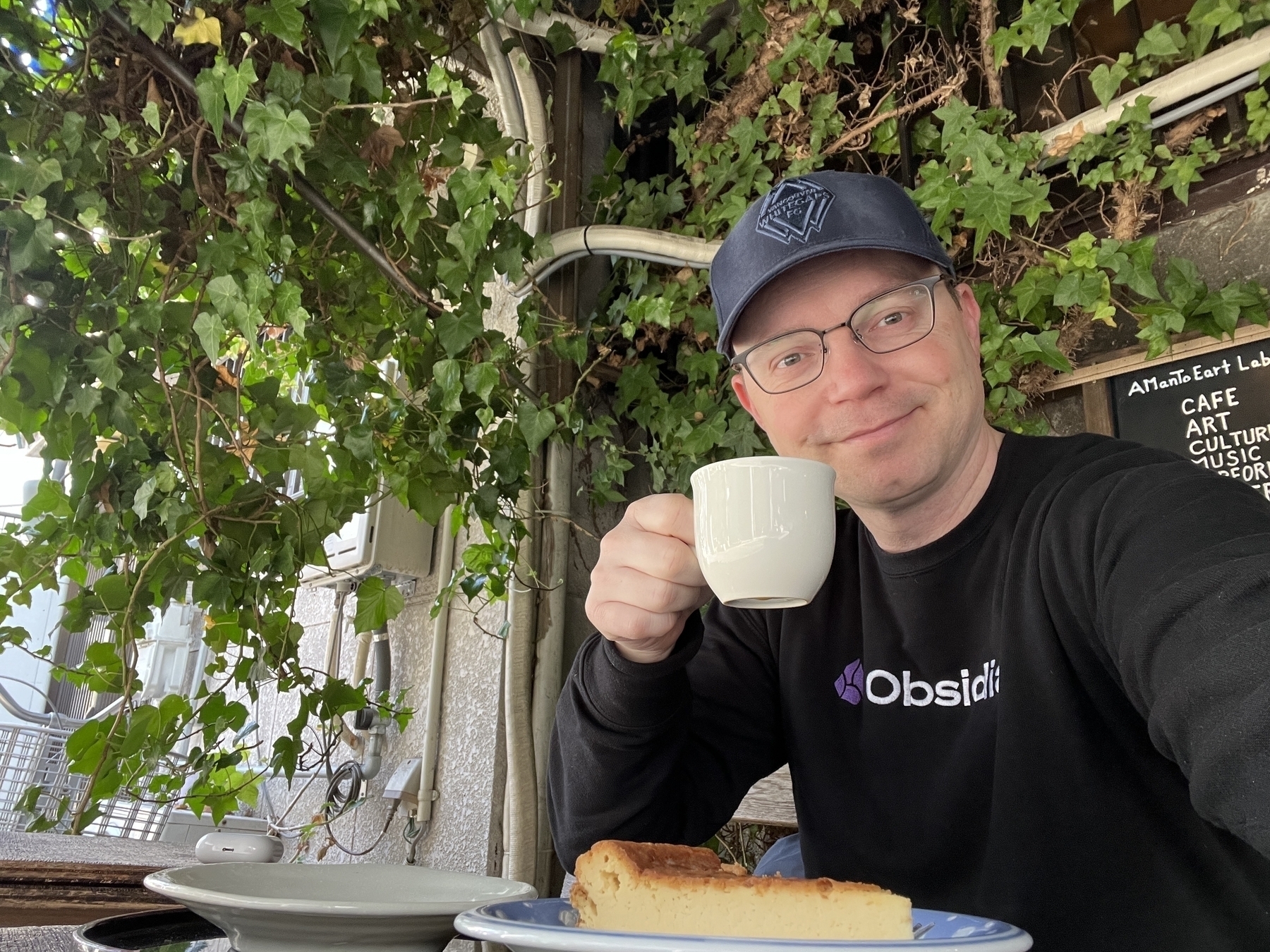 Chad sits outside of a cafe under a trestle covered in vines with a cup of coffee and slab of cheesecake