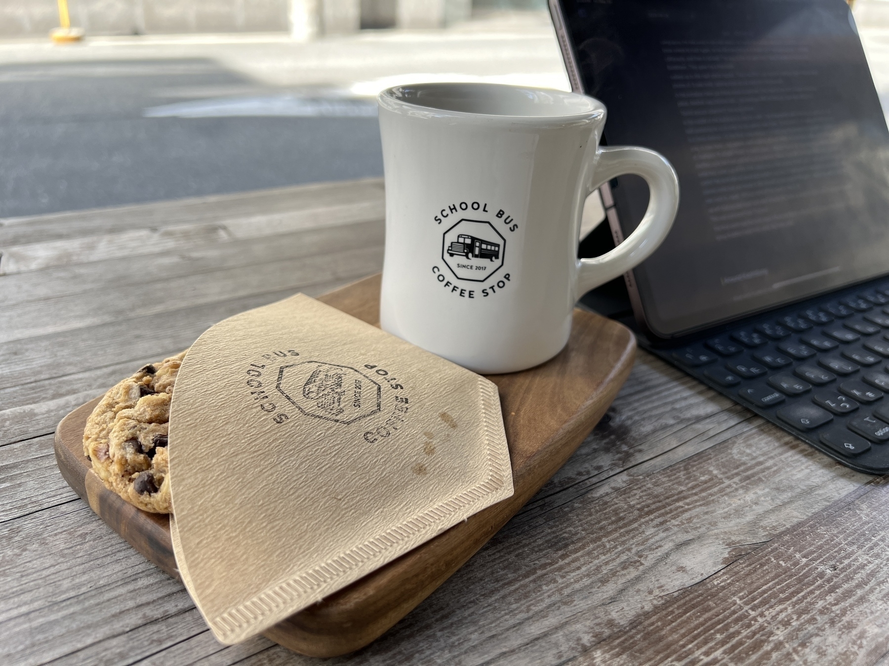 A cookie and cup of coffee beside an iPad on a table outside. The cookie is served in a coffee filter stamped with the School Bus Coffee Stop logo. 