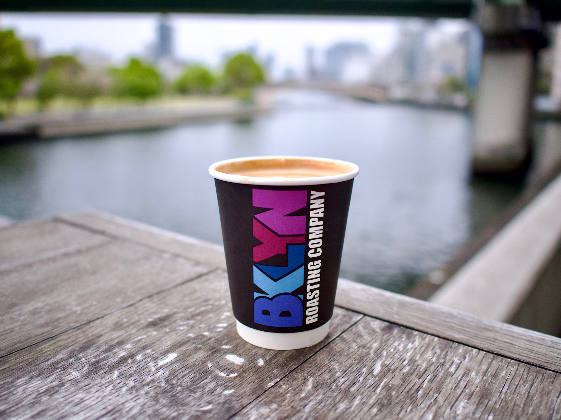 Paper Coffee cup with Brooklyn Roasters branding on a weather worn wooden table with a river in the background