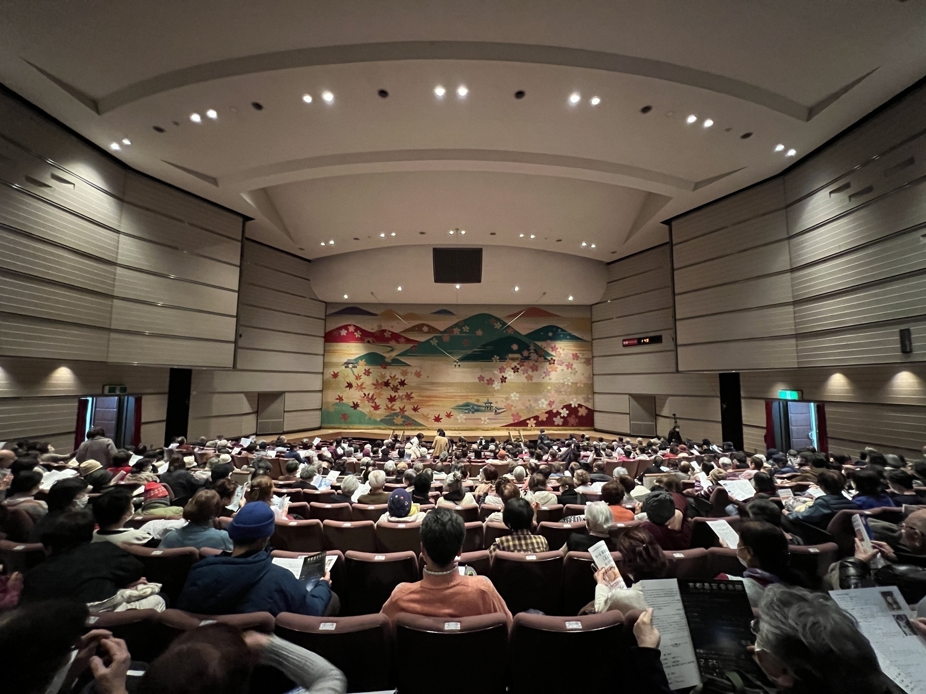 Theatre from the back. The brocade curtain displaying Biwako scenery is down and people are seated waiting. 