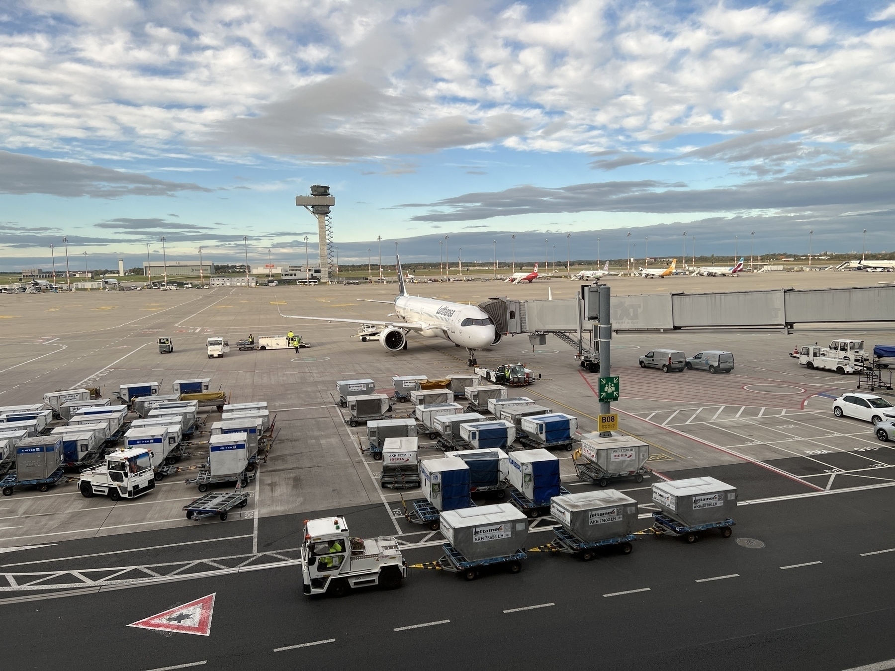 A Lufthansa airplane on the tarmac about to load passengers. The control tower is in the background. The morning sky is mottled with clouds