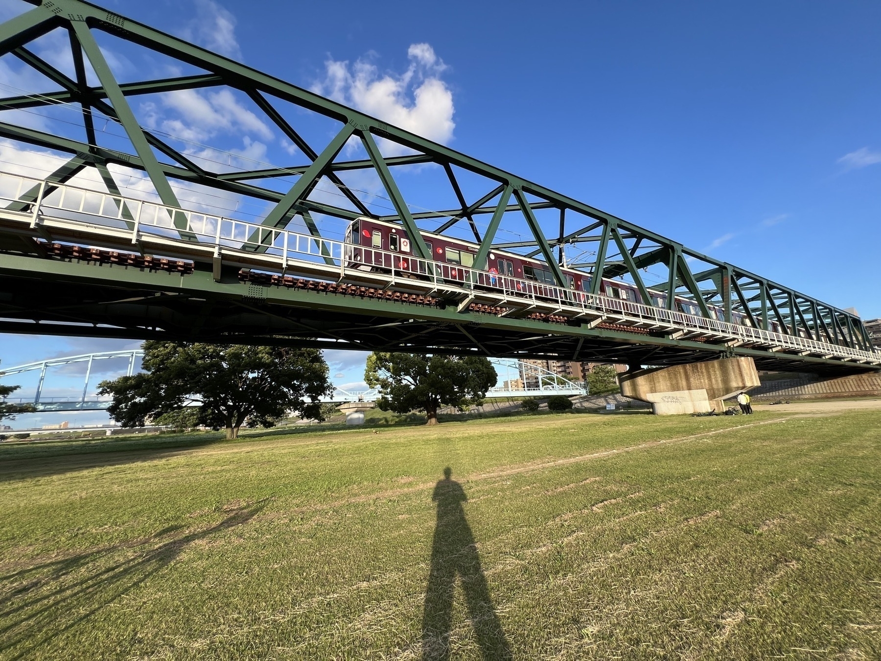 train with Myaku Myaku, the 2025 Osaka Expo mascot, passes over head on a tran bridge. Chad takes a photo from a grassy field below. The scene is lit by a low sun from behind, casting Chad (and his bike's) shadow across the grass