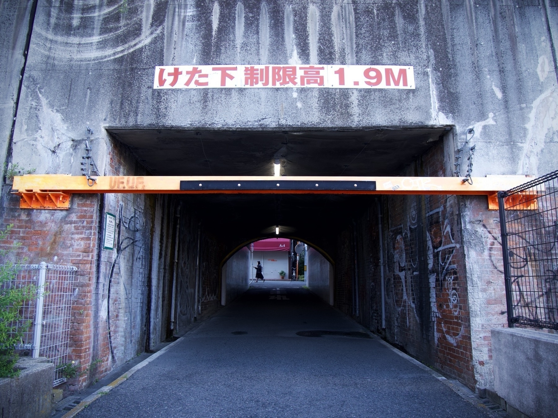 dark underpass under railway tracks during the day. The underpass is initally squared off, but then partway there is an arch from a previous period. On the far side a woman walks to the left. Above is a sign that says けた下制限高1.9M indicating the max height is 1.9m
