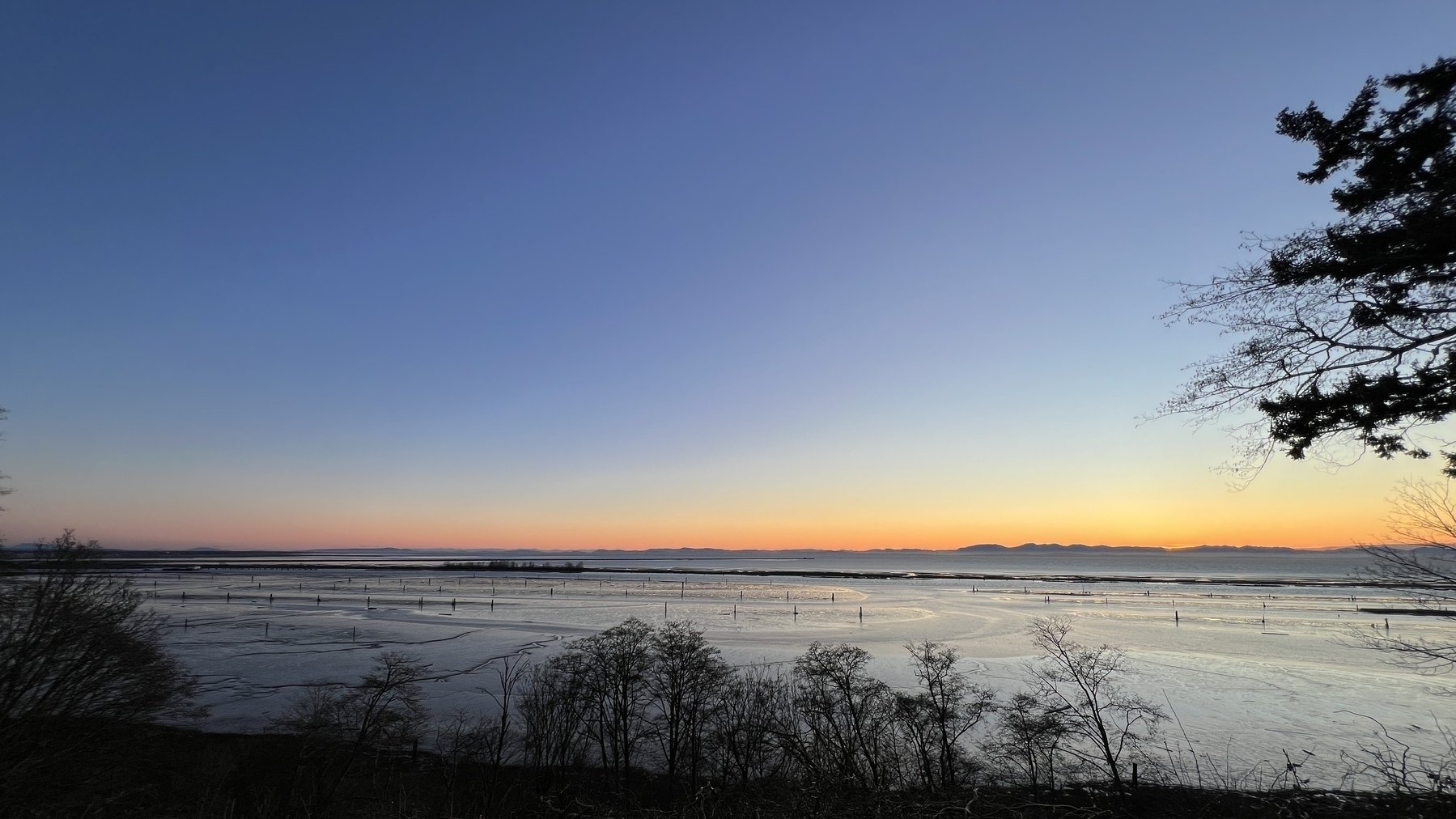 Dusk. From a high cliff the shallow waters of the ocean. In the far distance is Vancouver Island, above which the sky is orange from sunset. Above the sky turns dark blue of evening 