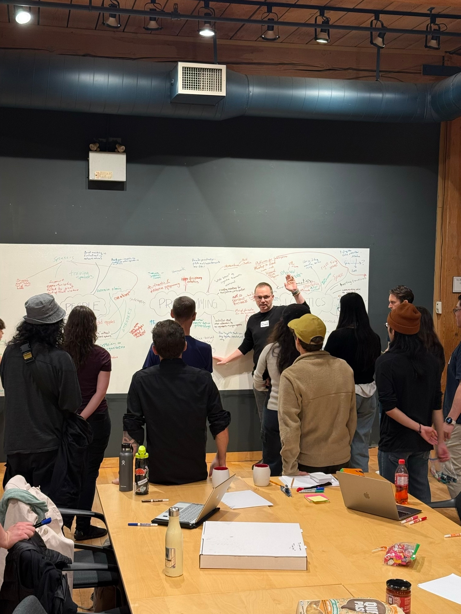 People crowd around a whiteboard with three brainstorming clouds around PEOPLE, PROGRAMMING, and LOGISTICS. Chad gesticulates wildly as usual