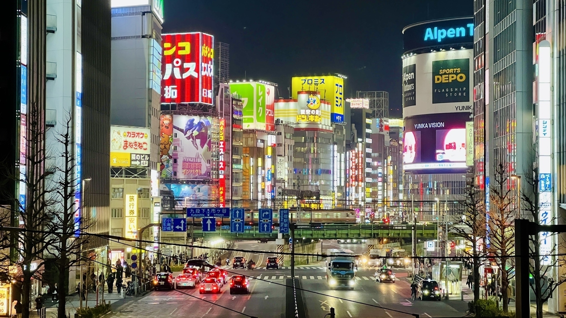 Zoom shot down a Shinjuku street as the Chuo train crosses a train bridge, behind which are many lit up signs