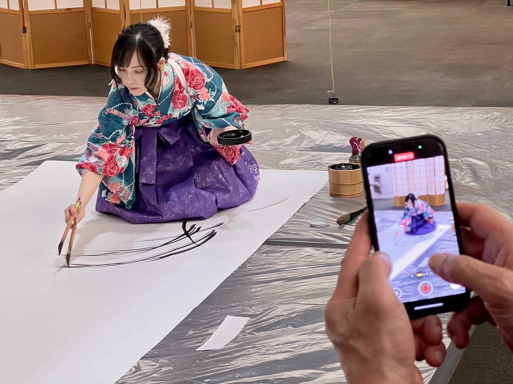 Woman in an ornate kimono kneeling on a large paper painting wheatgrass. In the foreground a person is videoing her