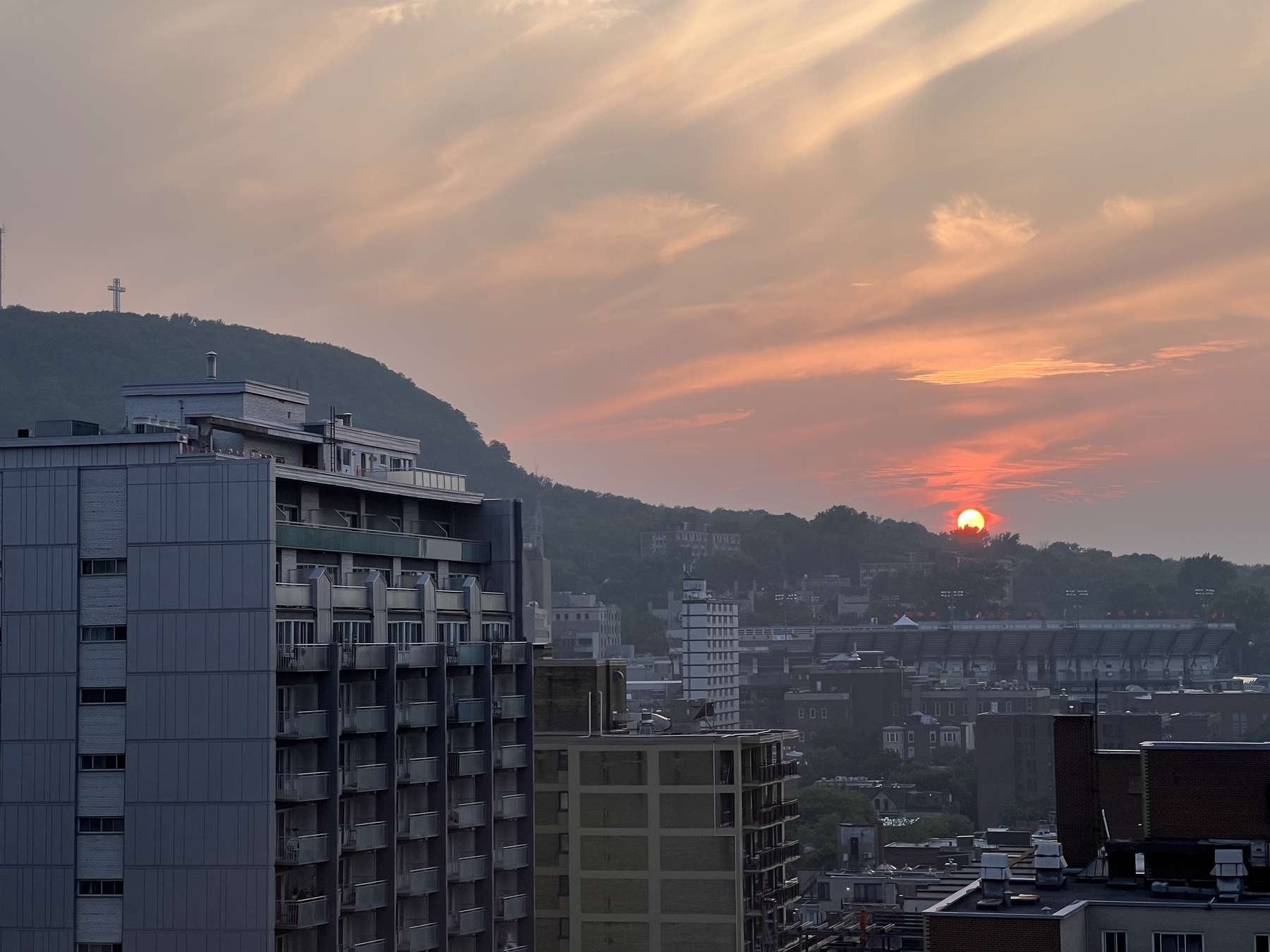 Orange sun in a smoky sky sinking behind the horizon. To the left is Mont Royal and the big cross looking down over the sun